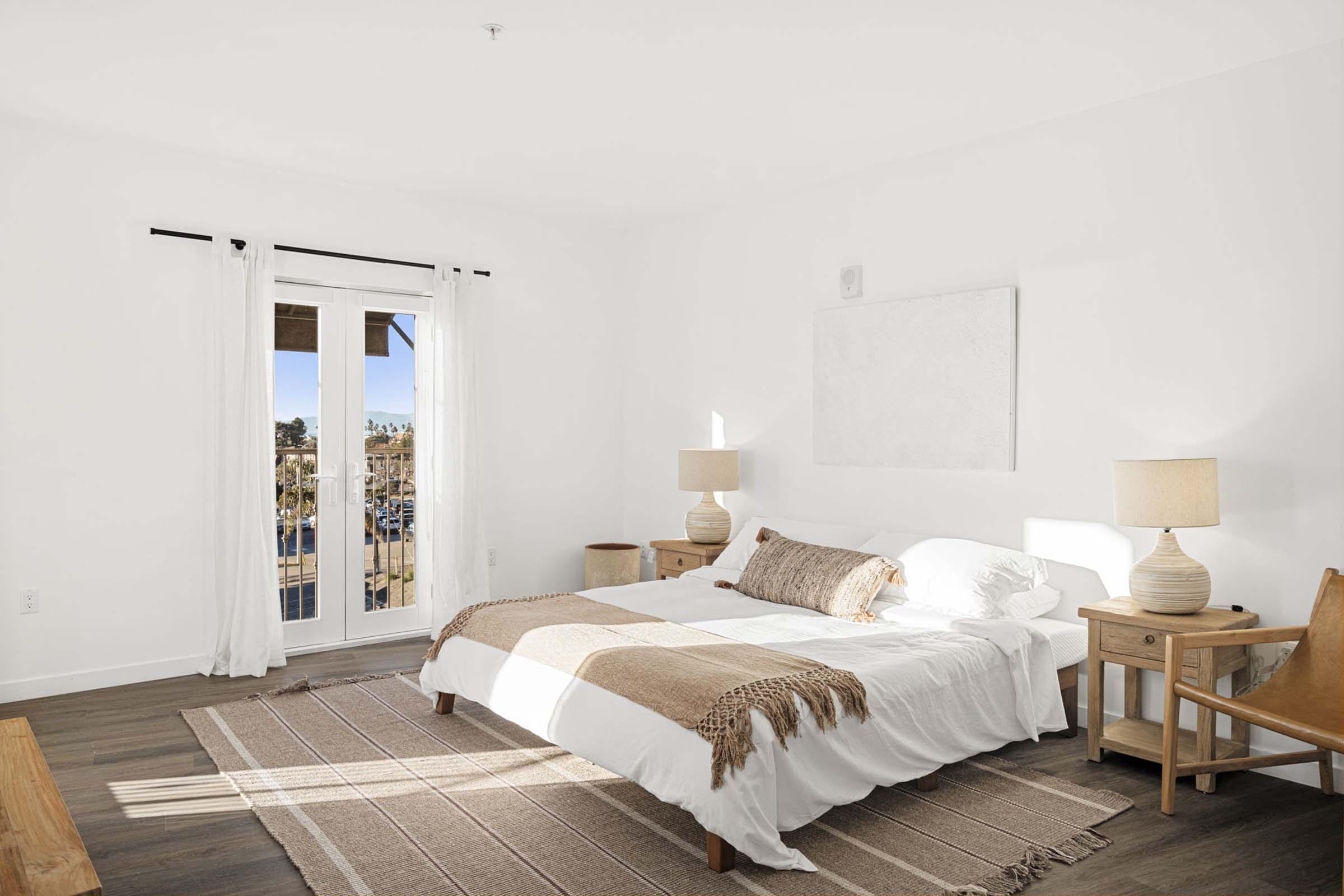A bright, minimalist bedroom featuring a white bedspread, wooden bedside tables with lamps, and a woven rug. Large windows let in natural light, enhancing the airy feel of the space. A small outdoor balcony is visible in the background, surrounded by greenery.
