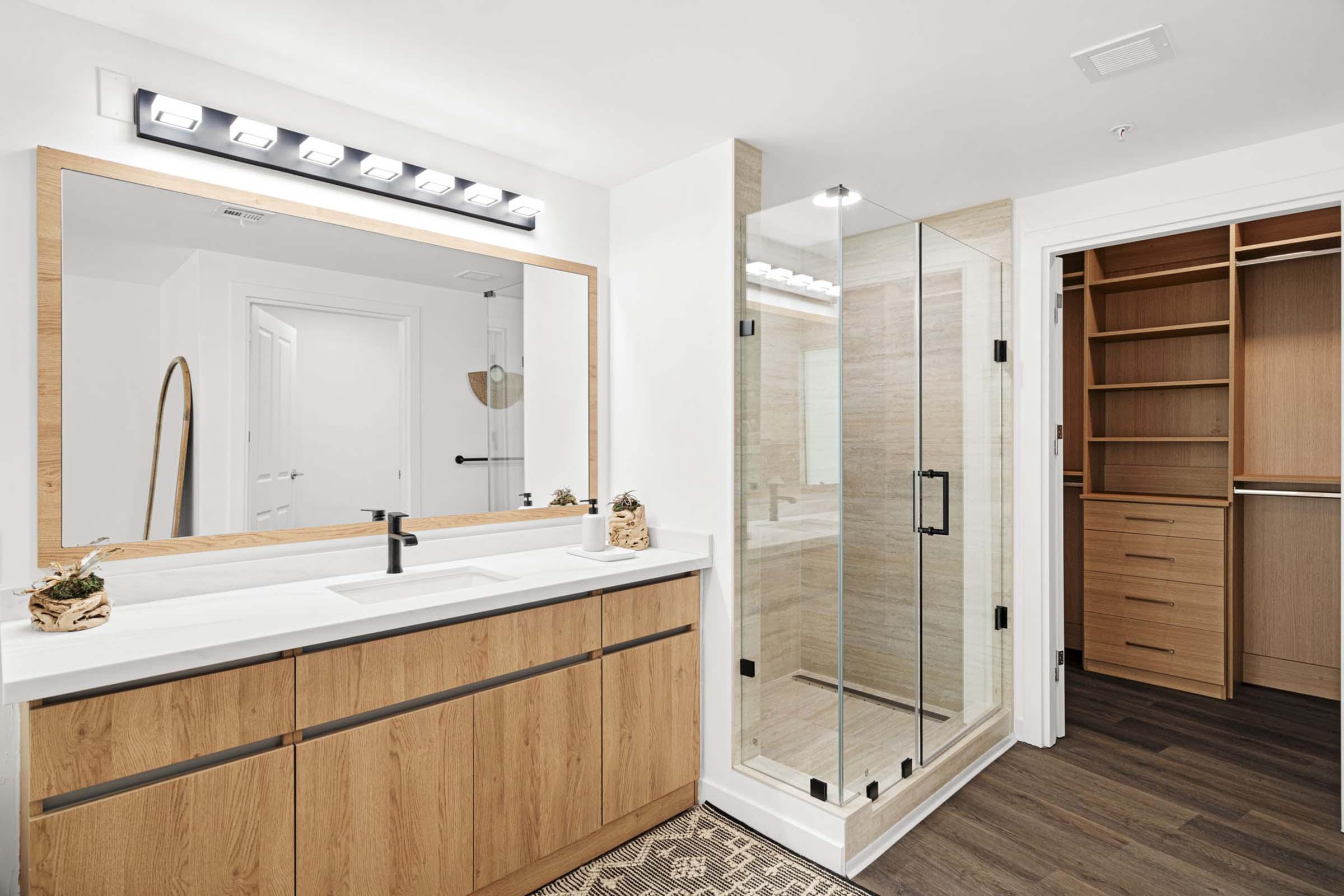 Modern bathroom featuring a minimalist design with a large mirror above a double sink vanity, wooden cabinetry, and a walk-in shower with glass doors. The room has soft lighting and a wooden shelving unit for storage, creating a sleek and inviting atmosphere.