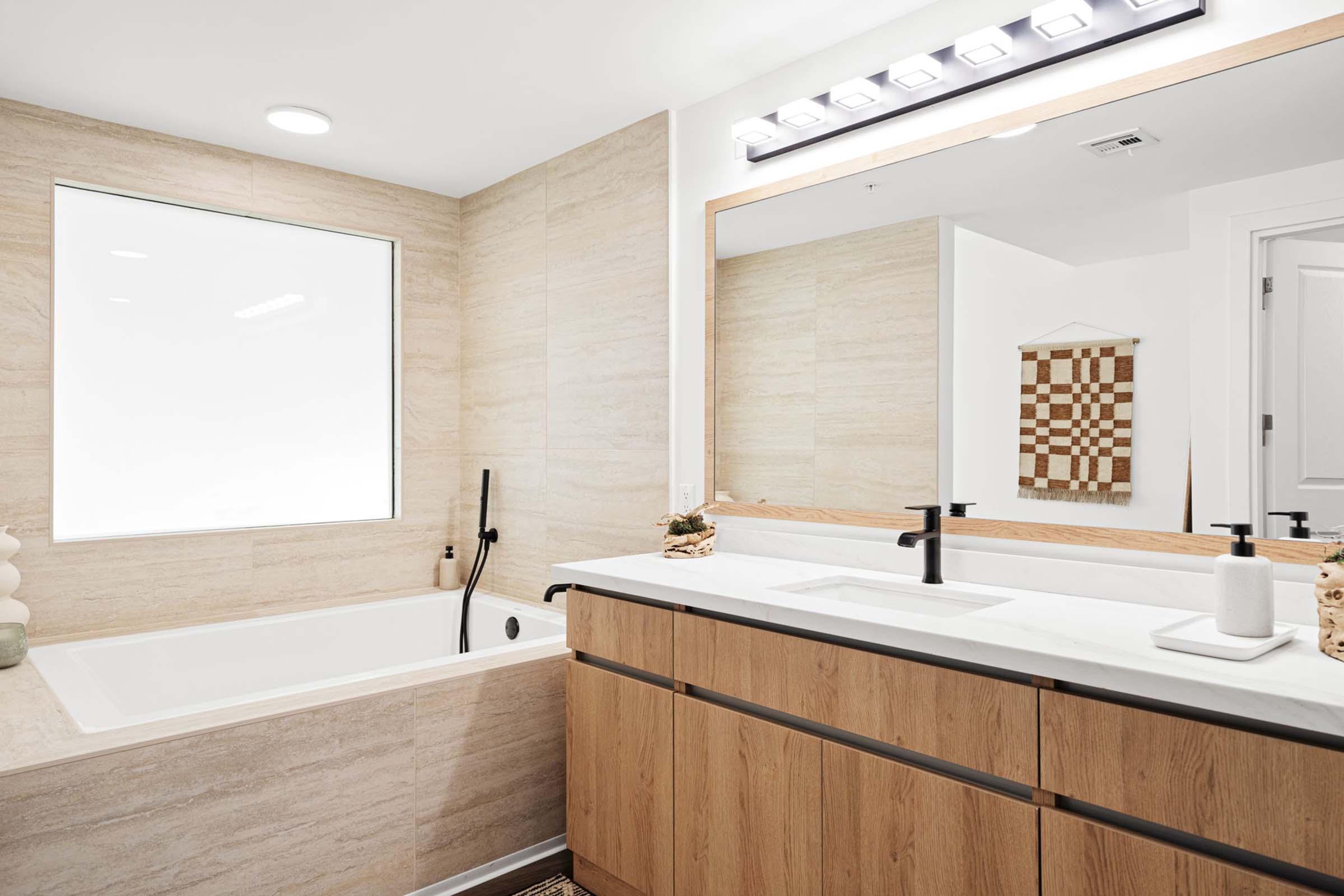 Modern bathroom featuring a sleek design with a large soaking tub, a double vanity with wooden cabinets, and a stylish mirror. Natural light streams in through a large window, complemented by neutral-toned walls and elegant fixtures. Decorative elements include a small woven wall hanging and minimal accessories.