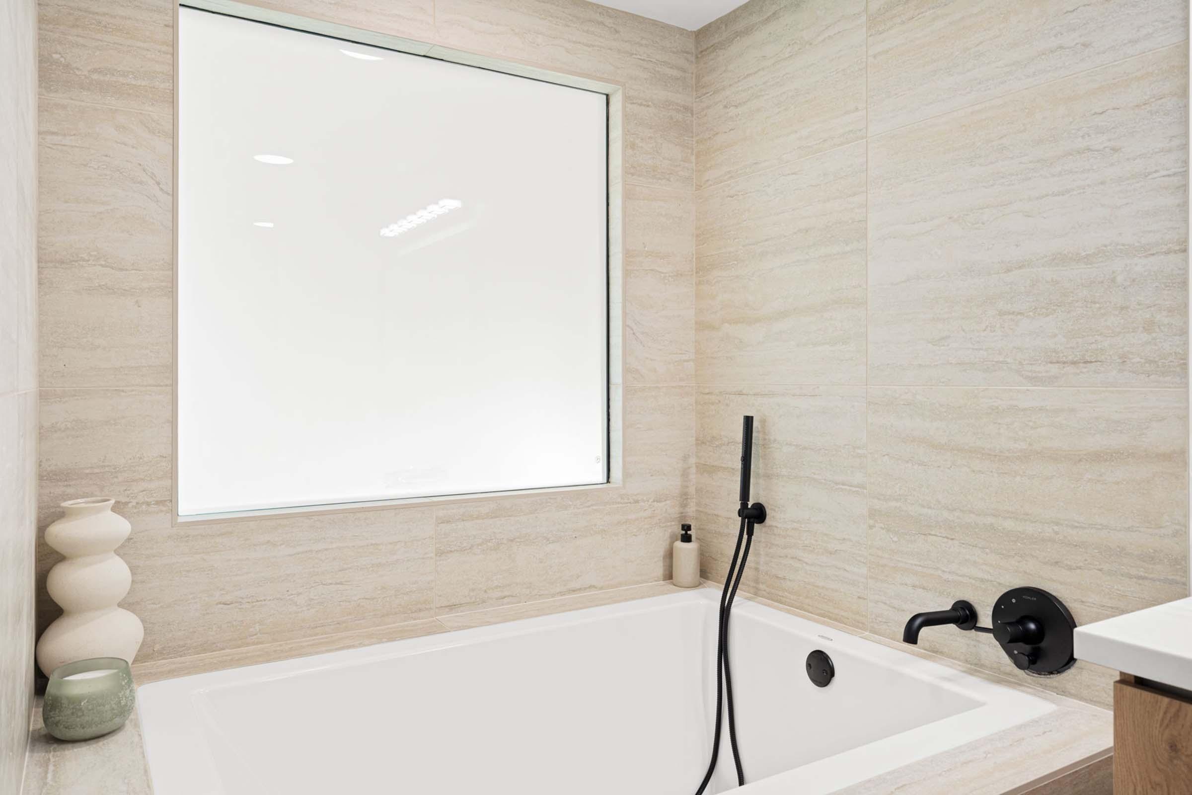 A modern bathroom featuring a white bathtub with a handheld shower fixture. The walls are adorned with light, textured tiles, and a large window provides ample natural light. A minimalist decorative vase sits beside the tub, contributing to the contemporary aesthetic.
