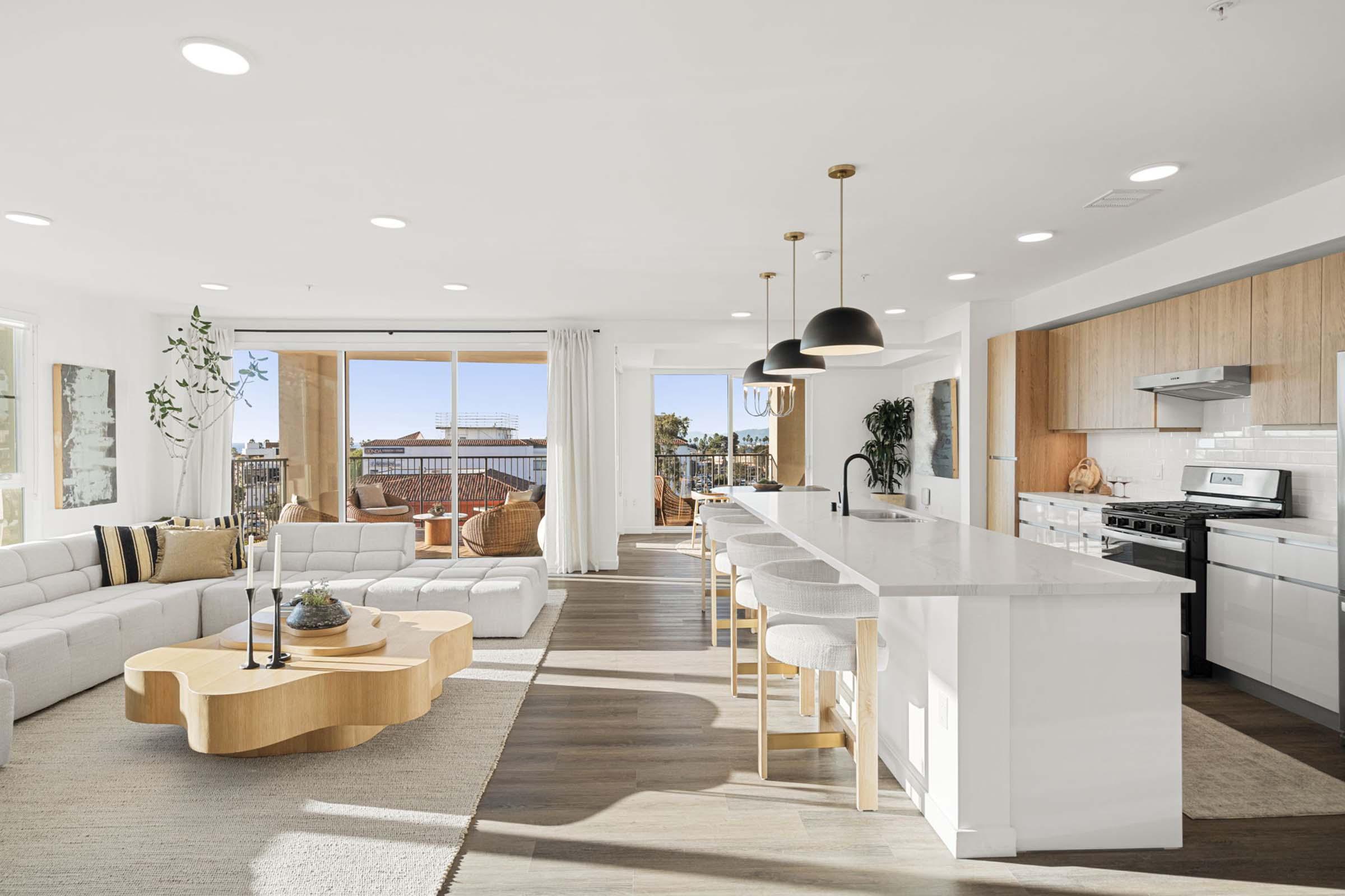 Spacious modern living area featuring a light-colored sofa, wooden coffee table, and an open kitchen with sleek countertops. Large windows bring in natural light, with a dining area visible. Stylish pendant lights hang above the kitchen island, creating a warm and inviting atmosphere.