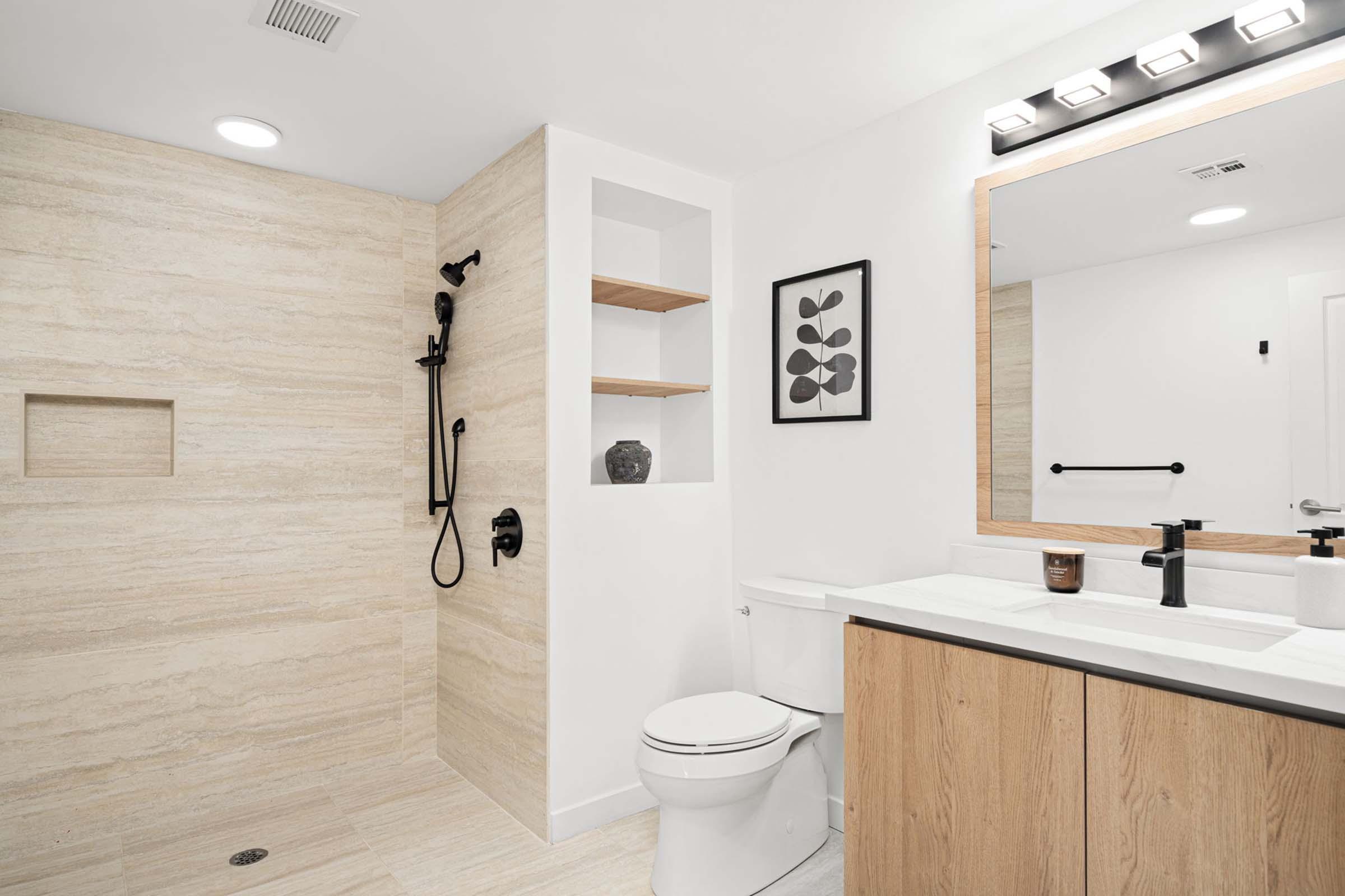 A modern bathroom featuring a walk-in shower with a black shower fixture, a wooden vanity with a sink, a white toilet, and minimalist decor including a framed artwork and open shelves. The room has neutral-colored walls and warm lighting for a contemporary look.