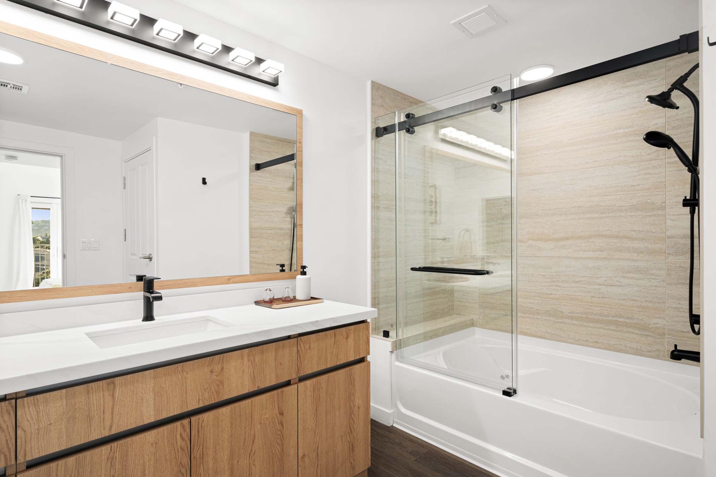 Modern bathroom featuring a glass shower enclosure and a bathtub. The sleek design includes a double vanity with wood cabinetry and a large mirror above. Soft lighting illuminates the space, and a window lets in natural light, enhancing the contemporary aesthetic.
