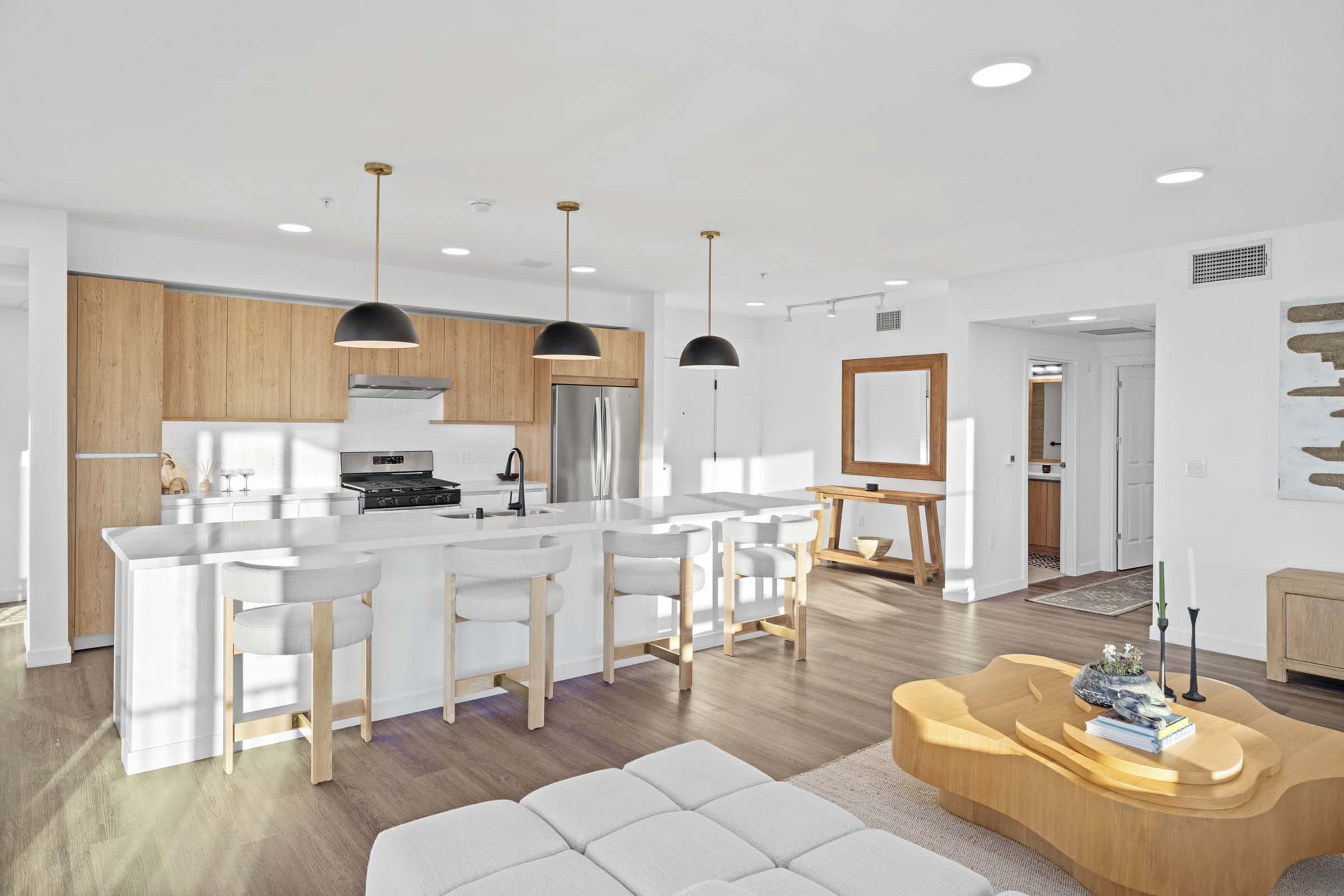 A modern kitchen and living area featuring a white countertop island with bar stools, wooden cabinetry, pendant lights, and stainless steel appliances. The open space includes a light-colored sofa, a wooden coffee table, and a decorative piece on a side table, with natural light streaming in from large windows.