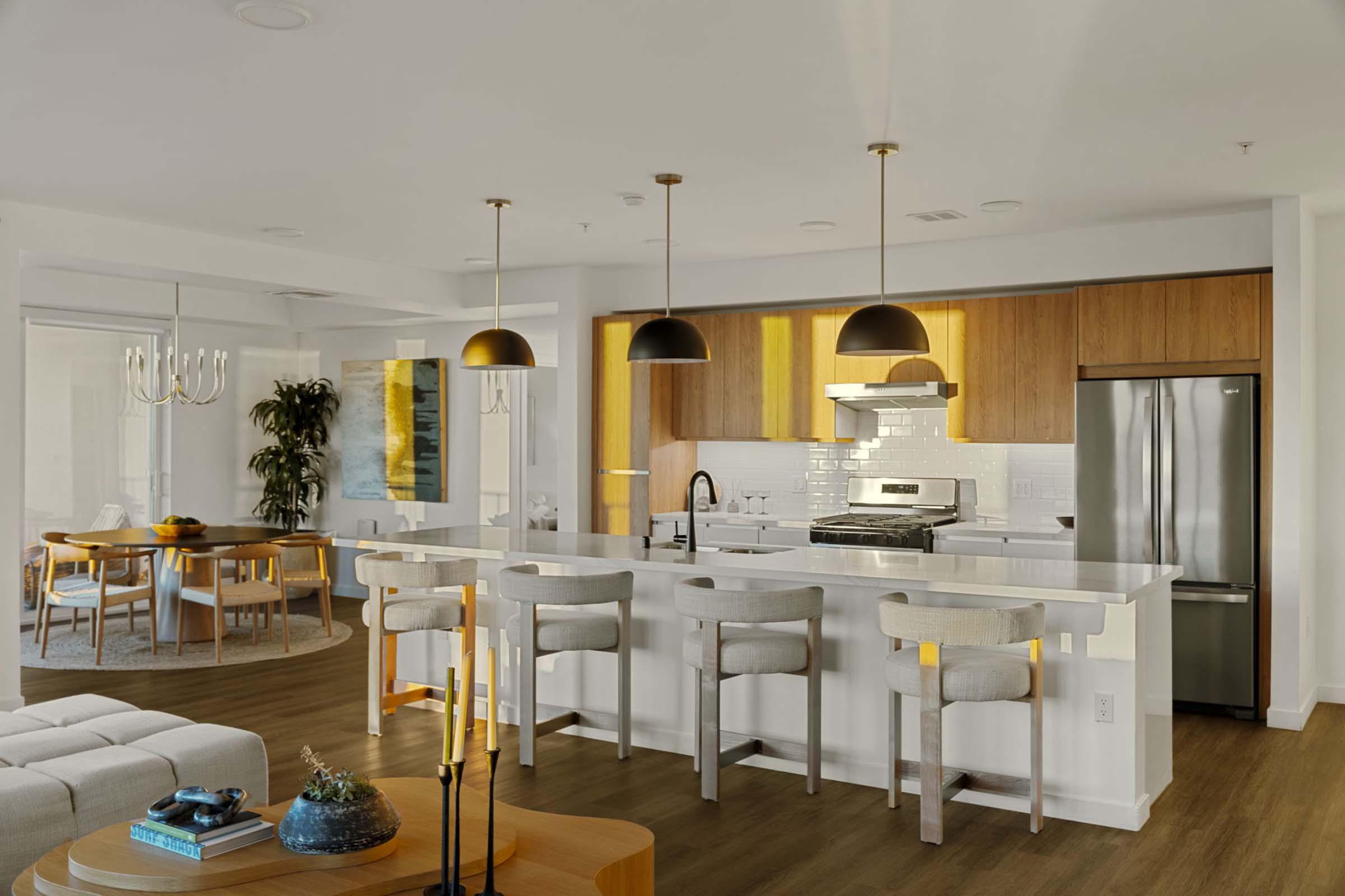 Modern open-concept kitchen featuring a large island with four stools, stainless steel appliances, and warm wood cabinetry. The dining area includes a round table with wooden chairs, and a cozy living space is visible in the background with a light color palette and contemporary decor.