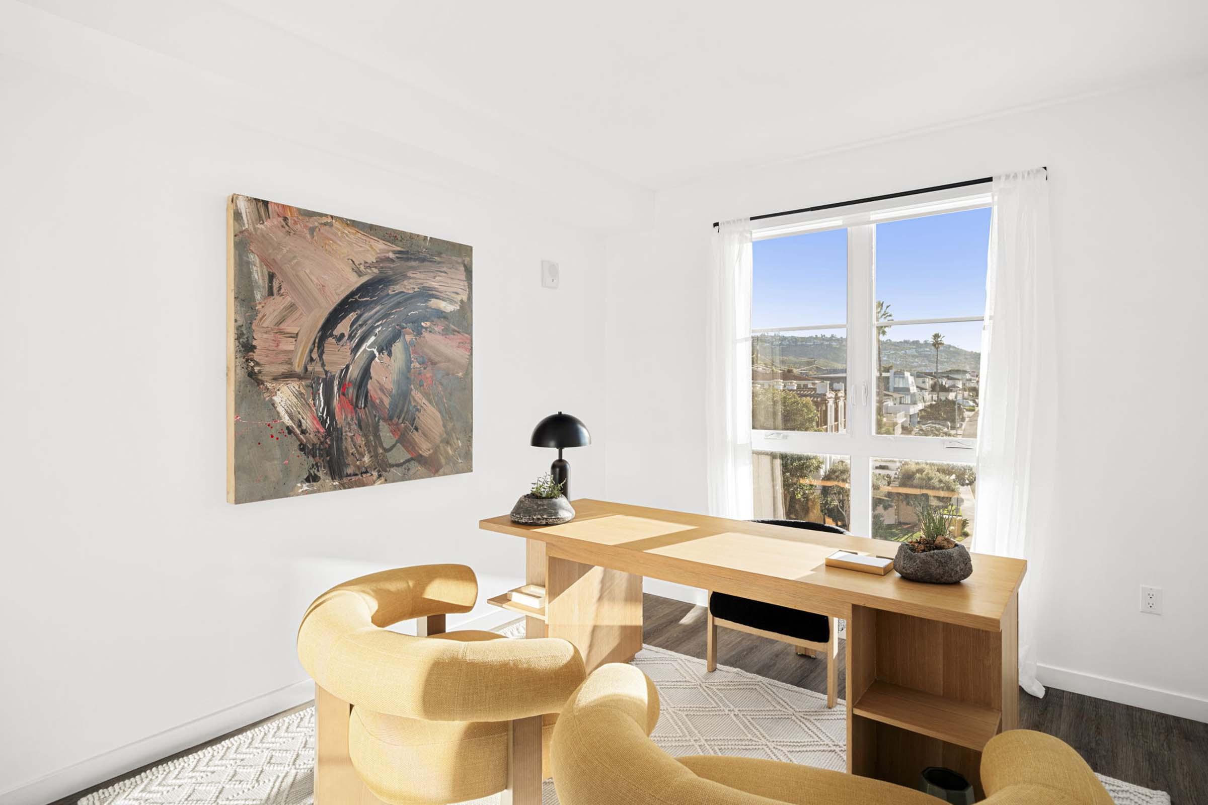 A bright, modern home office featuring a wooden desk with a lamp and decorative items, two curved yellow chairs, and a large abstract painting on the wall. A window offers a view of the outdoors, letting in natural light, and white walls create a spacious feel.