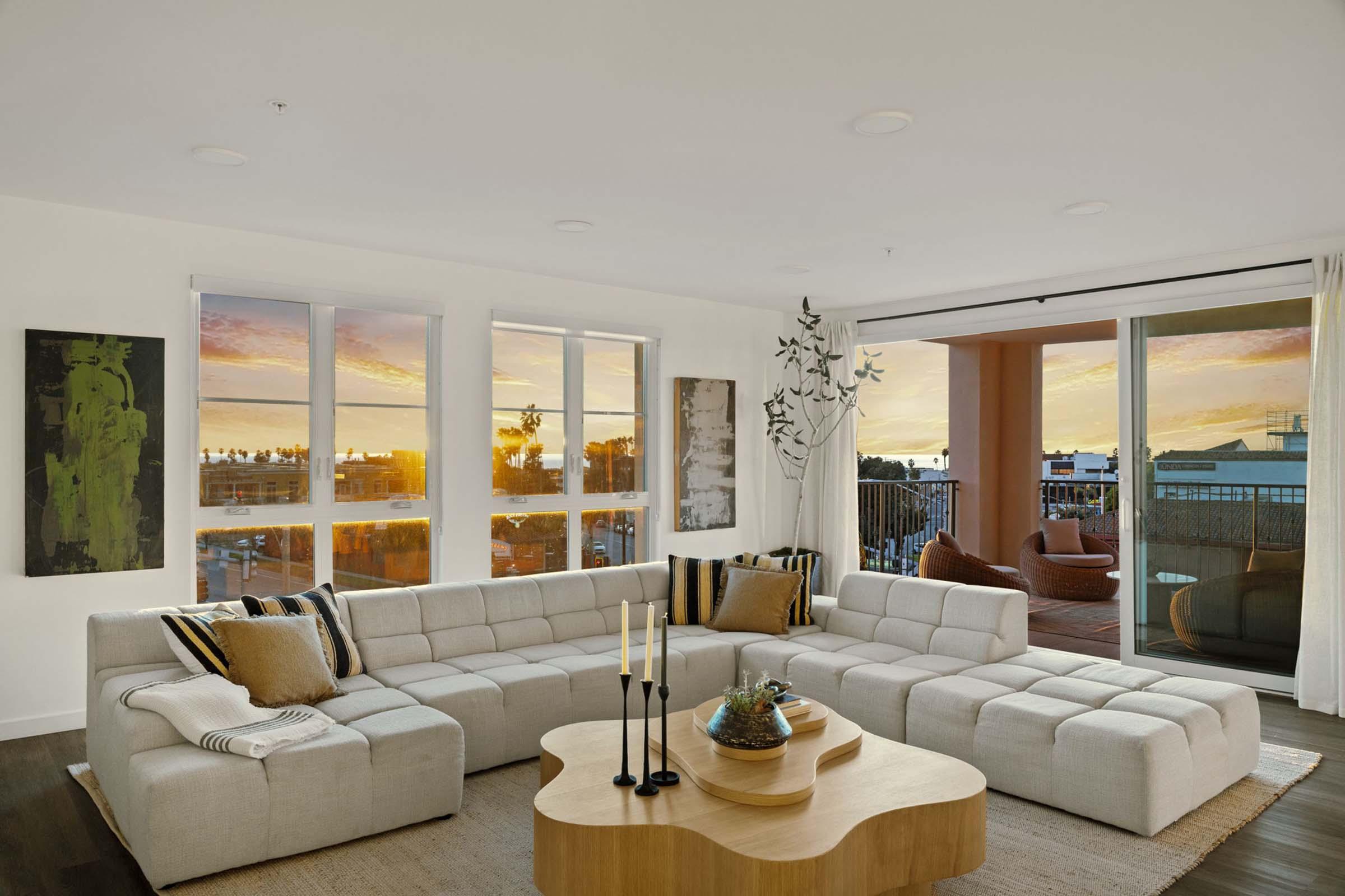 A modern living room featuring a light-colored sectional sofa, a wooden coffee table, and decorative accents. Large windows offer a view of a sunset outside, illuminating the space. The walls are adorned with abstract art, and there are green plants adding a touch of nature to the interior design.