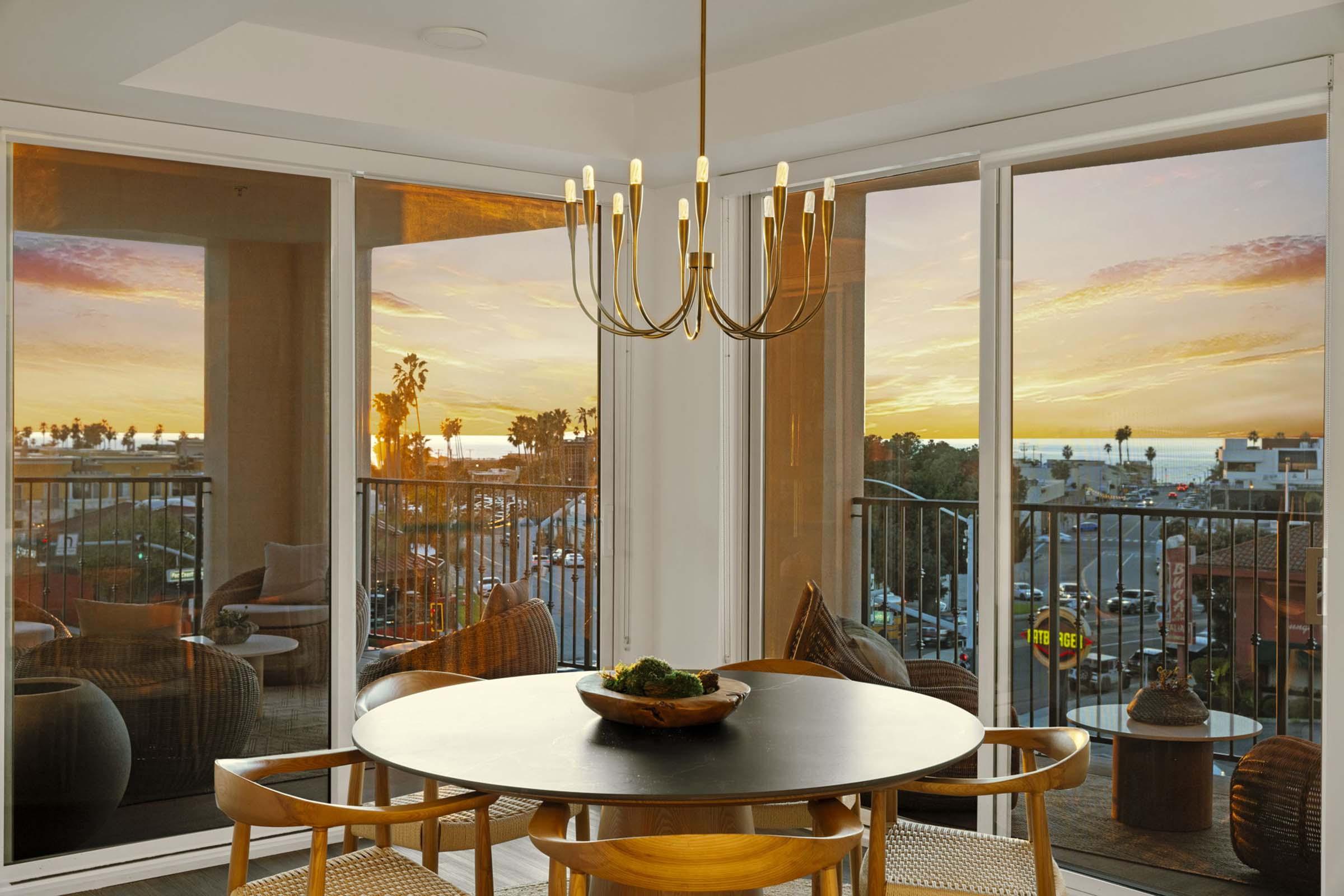 A modern dining area featuring a round black table with a wooden bowl centerpiece. Behind, large glass windows showcase a vibrant sunset over the ocean, with palm trees and city life visible. The space is warmly lit by a contemporary chandelier, creating an inviting atmosphere.