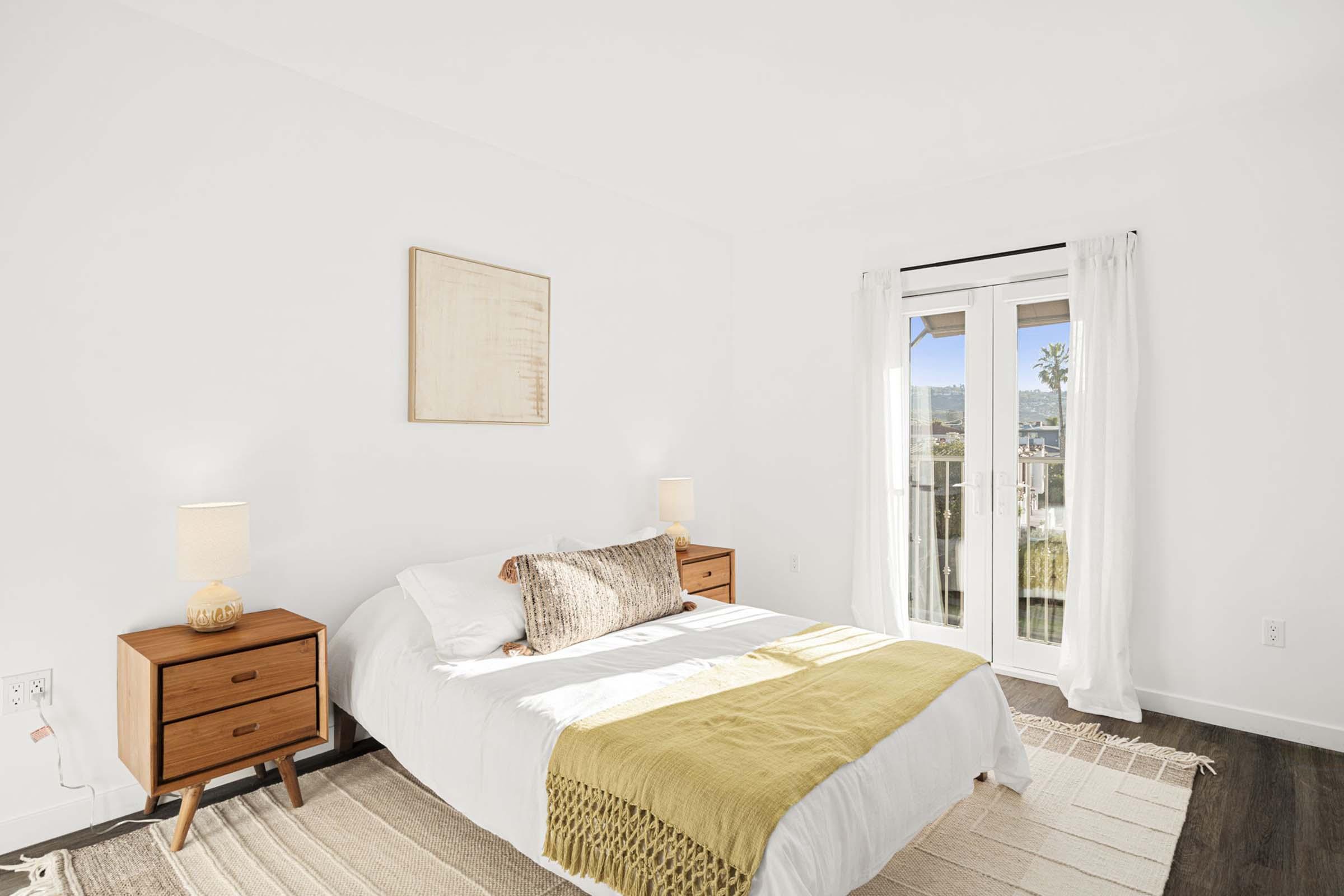 A cozy and bright bedroom featuring a queen-sized bed with a white duvet, decorative pillows, and a mustard-colored throw. There are two wooden nightstands with lamps on either side, and large windows with sheer white curtains that allow natural light to illuminate the space. A rug adds warmth to the wooden floor.