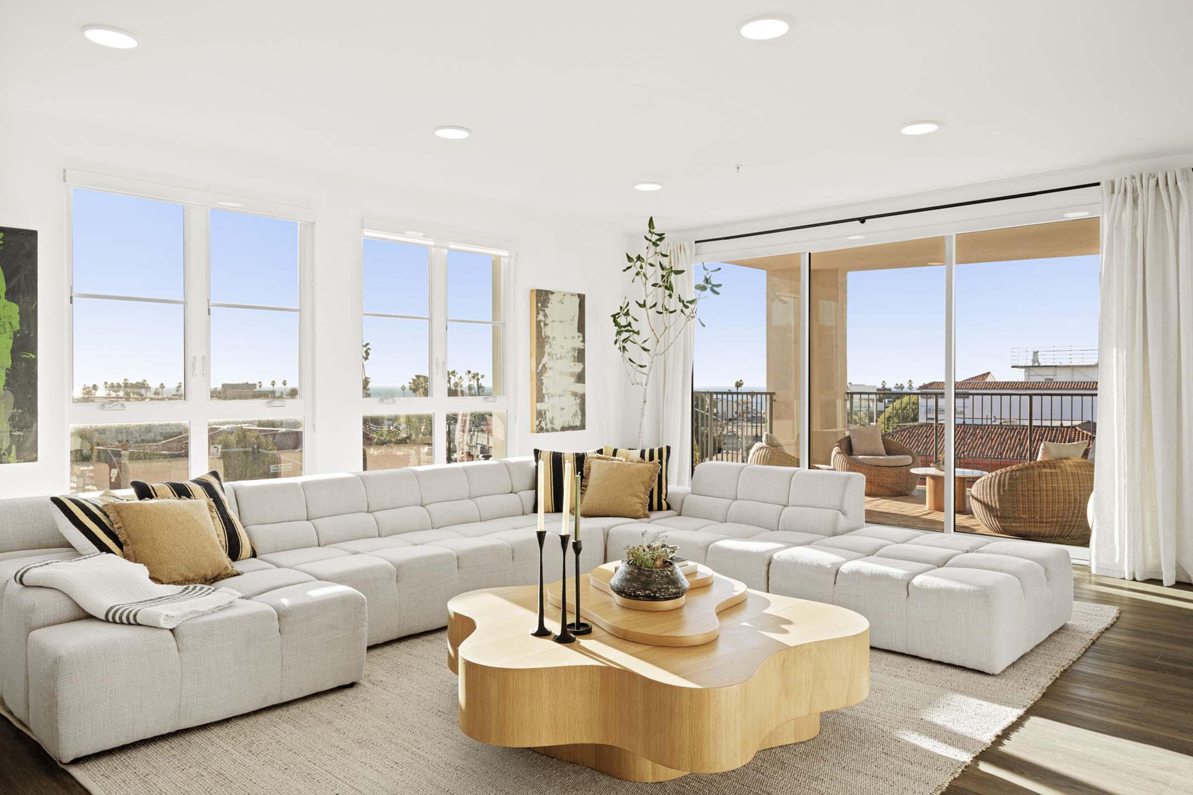 A modern living room featuring a large light-colored sectional sofa, a wooden coffee table with a unique shape, and large windows that provide natural light and a view of an outdoor balcony with seating. Decor includes minimalistic artwork and greenery. The overall aesthetic is bright and inviting.
