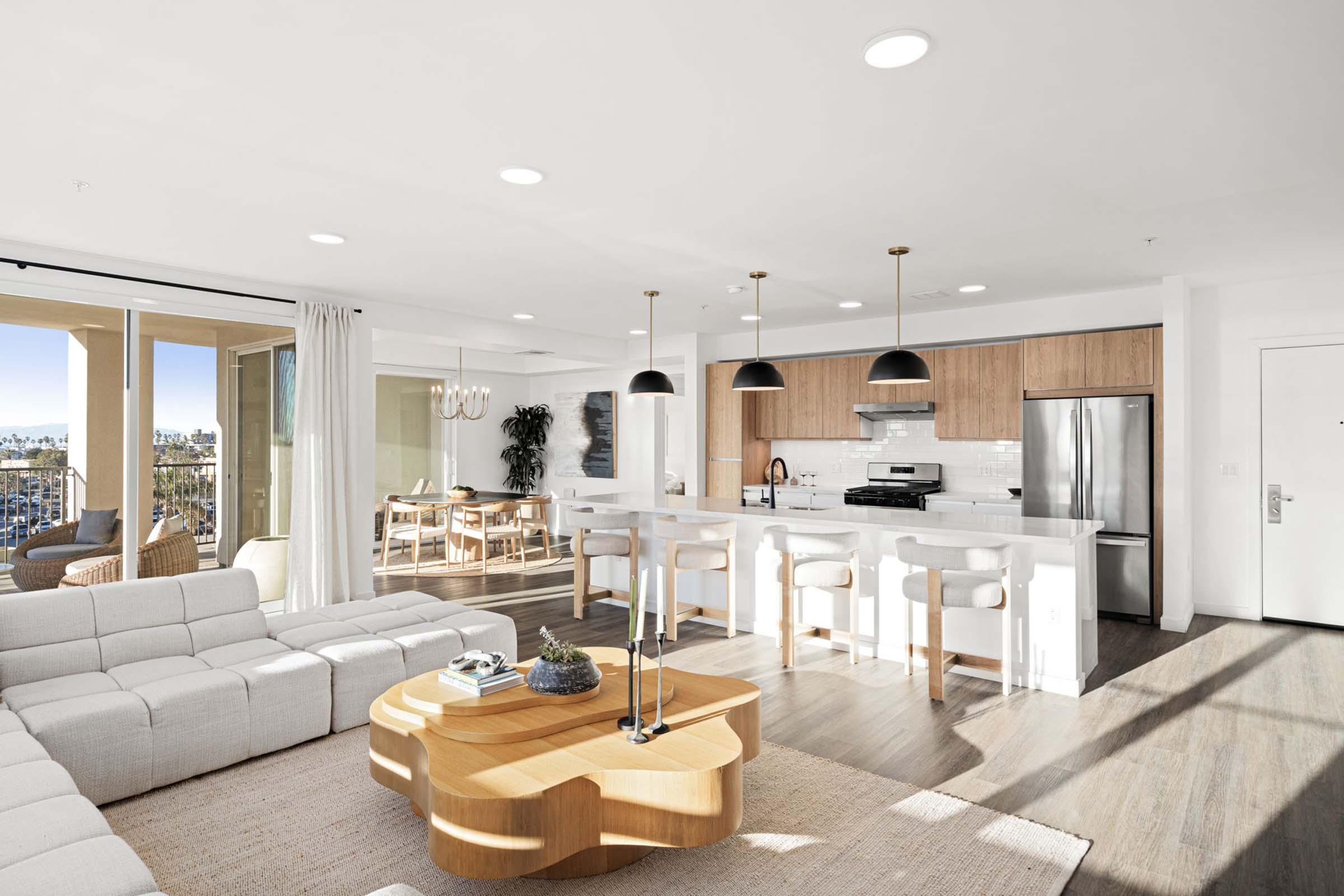 Spacious modern living room and kitchen with light wood accents, featuring a plush white sectional sofa, a stylish wood coffee table, and sleek pendant lighting. Large windows offer views of a sunny outdoor area, creating a bright and inviting atmosphere.