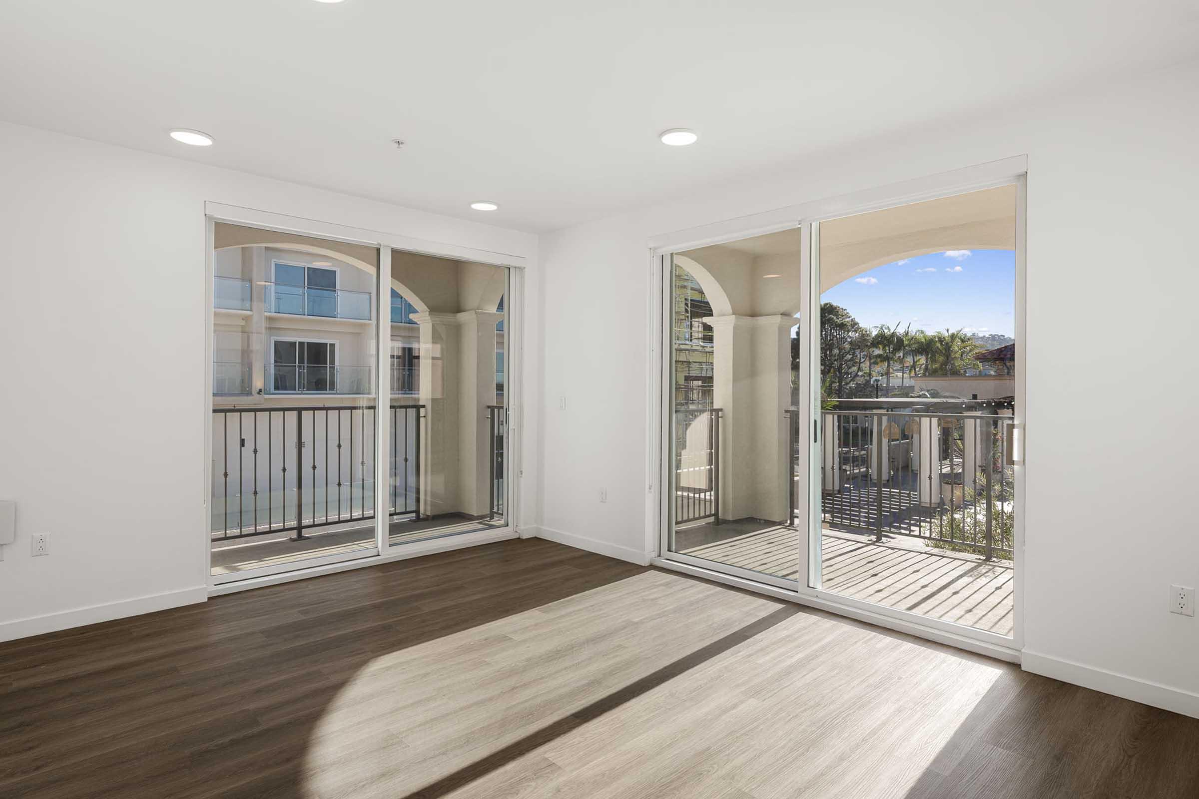 Bright, empty room with large sliding glass doors leading to a balcony. The floor is wood-like laminate, and the walls are painted white. Natural light pours in, creating an airy atmosphere. There are palm trees visible outside, suggesting a warm climate.