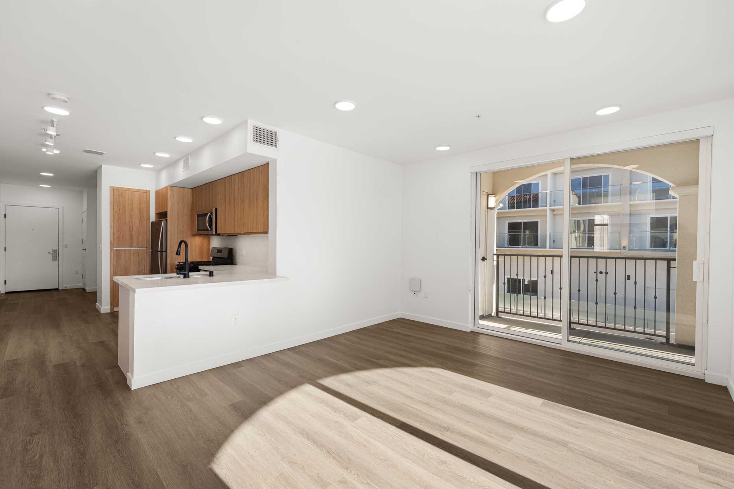 A bright, modern apartment interior featuring an open floor plan. The kitchen is equipped with wooden cabinetry, stainless steel appliances, and a large island. Sunlight streams through sliding glass doors that lead to a balcony, enhancing the spacious living area with light hardwood flooring.