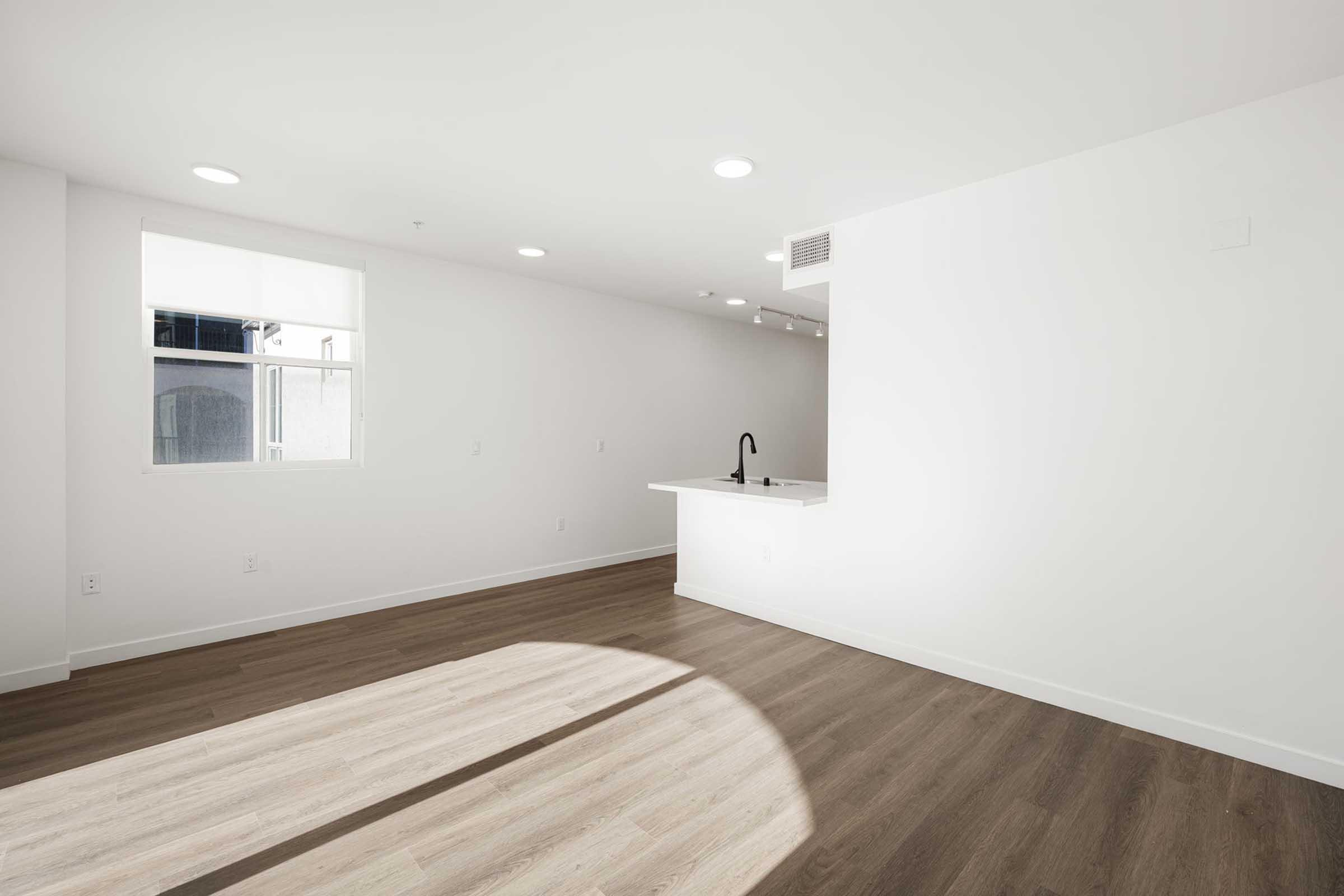 A spacious, well-lit room with white walls and a large window allowing natural light. The floor features light wood laminate, and there is a small kitchen area with a sleek black faucet on a white counter. The overall design is modern and minimalistic.