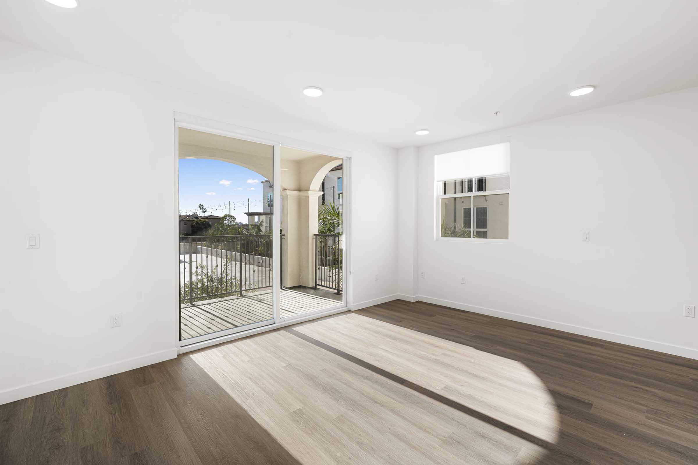 Bright, minimalist room with large glass sliding doors leading to a balcony. The walls are white and the floor features a mix of light and dark wood tones. Natural light fills the space, highlighting the open layout and modern design, with a view of greenery outside.
