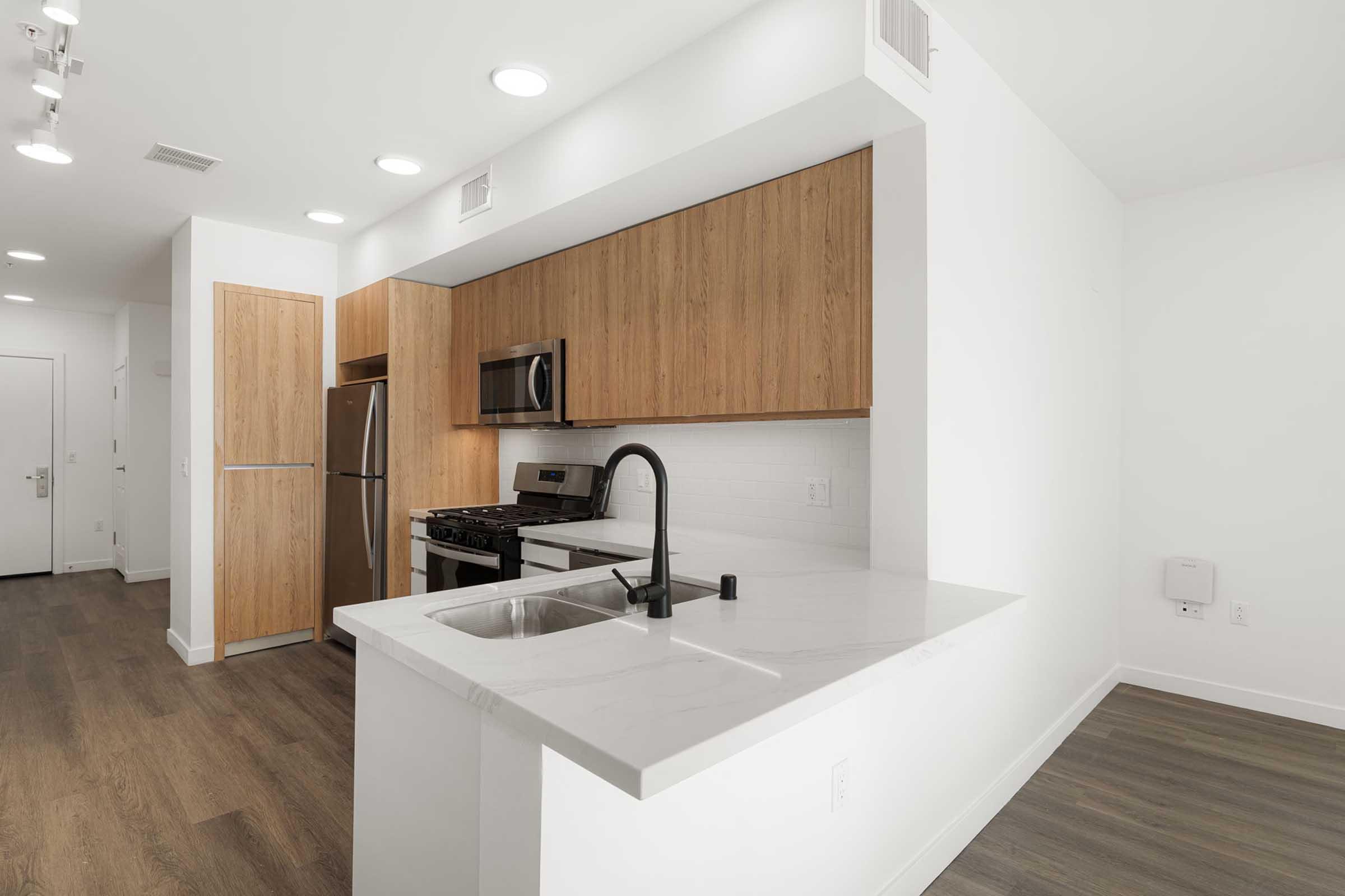 Modern kitchen featuring wooden cabinetry, stainless steel appliances, and a sleek marble countertop. The kitchen includes a sink with a black faucet, a gas stove, and a microwave. The open layout allows for a bright and spacious living area with light-colored walls and hardwood flooring.