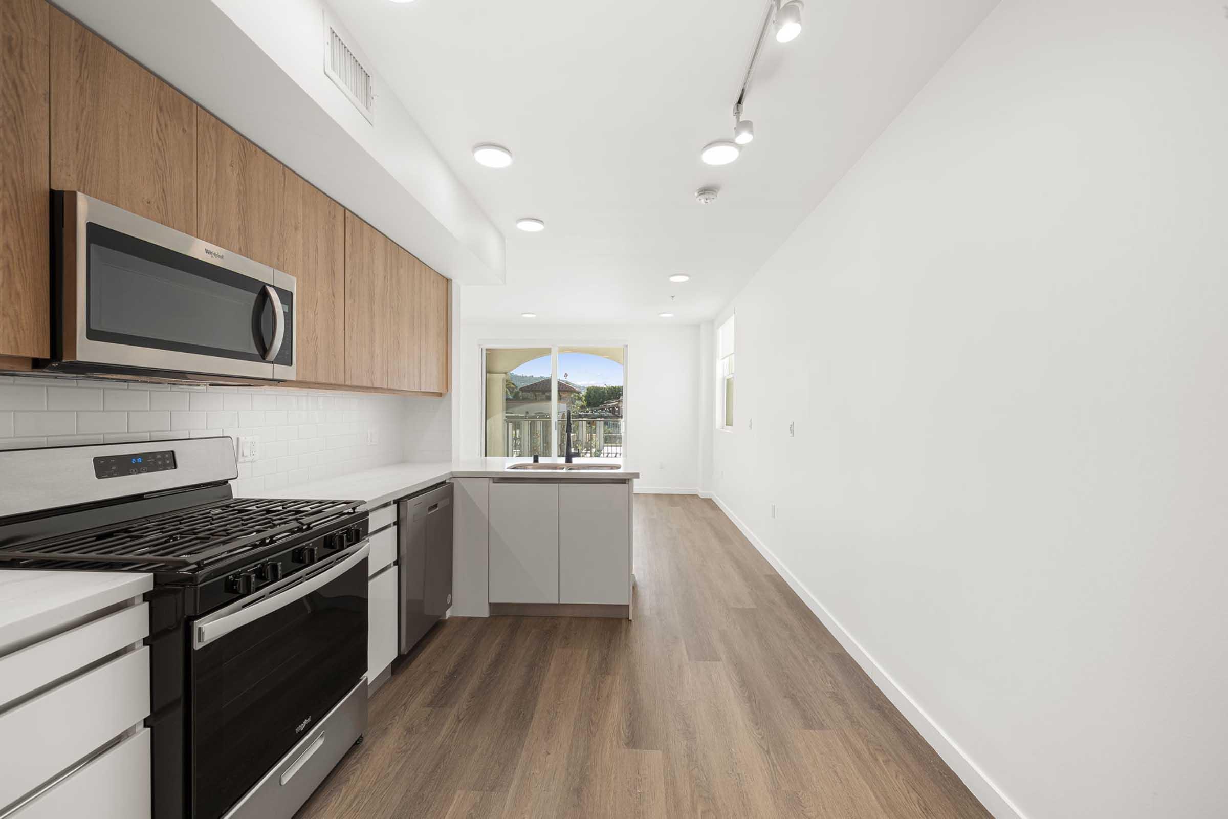 Modern kitchen featuring stainless steel appliances, including a gas stove and microwave, with light wood cabinetry. The space has a clean, minimalist design with white walls and wooden floors, and a large window allowing natural light to fill the room.