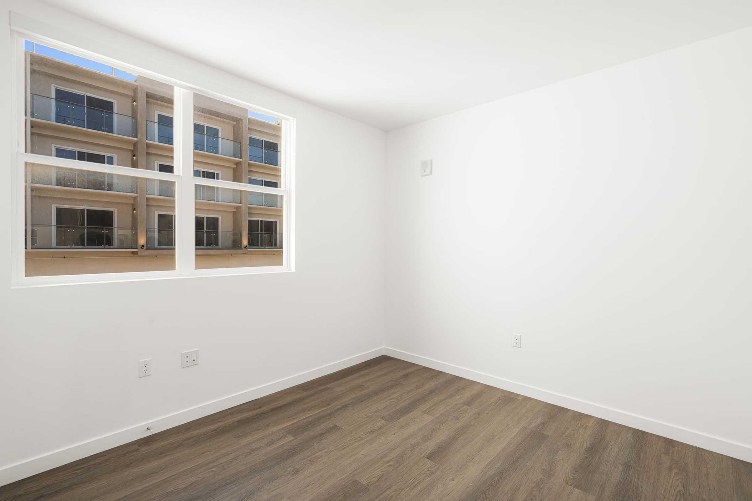 A bright, empty room with white walls and a window showcasing a nearby building. The flooring is light wood. Natural light floods the space, creating a clean and airy ambiance.