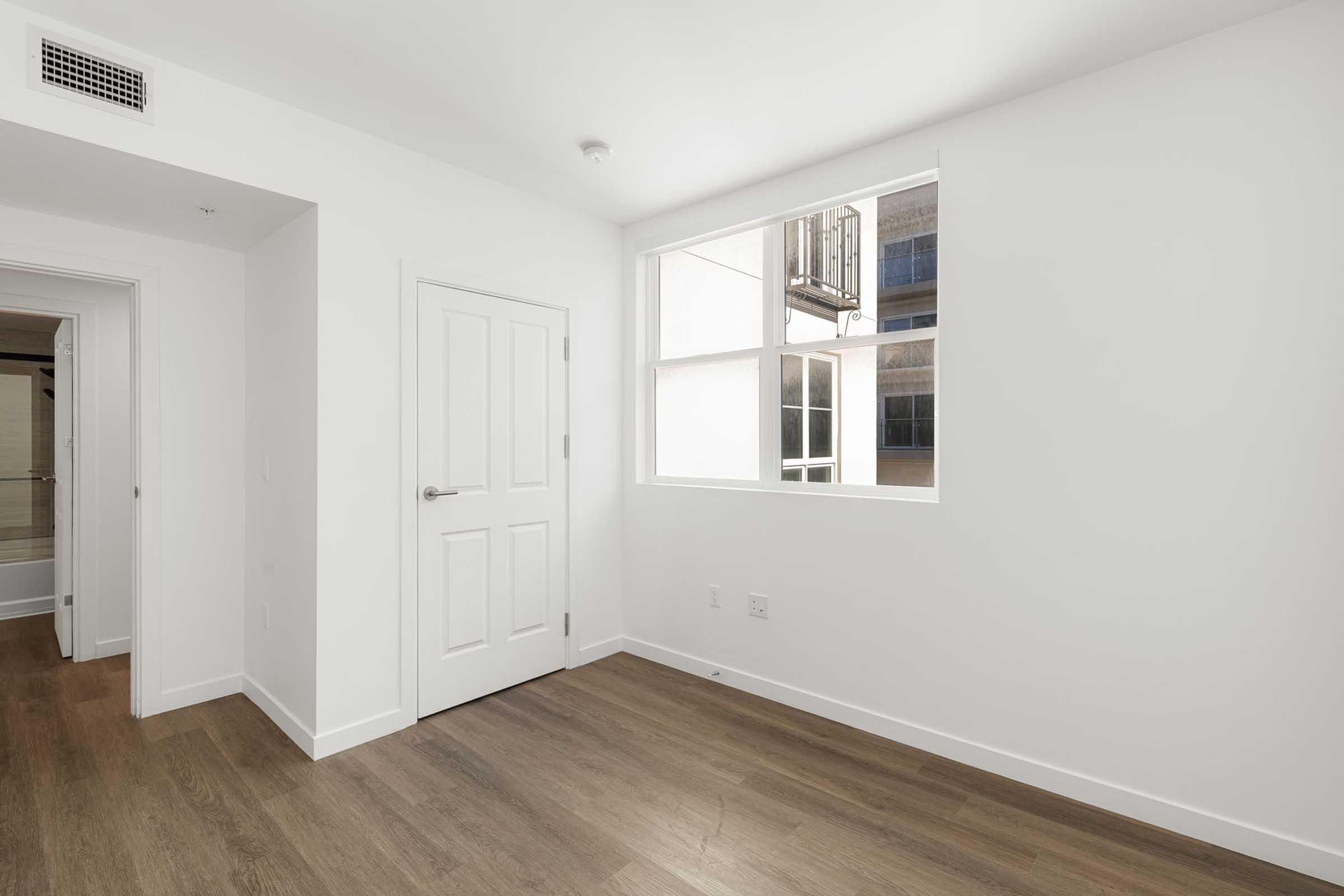 Bright, empty room with white walls and a wooden floor. A large window lets in natural light, and there is a closed white door on one wall. Visible is a doorway leading to another room in the background. The overall feel is clean and modern.