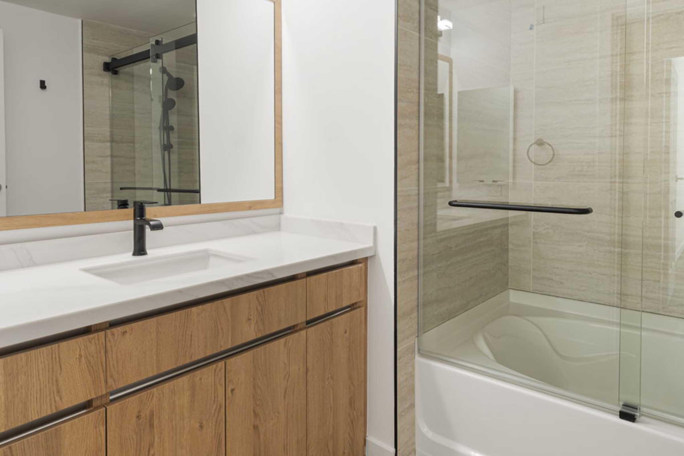 Modern bathroom featuring a wooden vanity with a white countertop and a single black faucet. A large, framed mirror hangs above the sink. The shower area is enclosed with glass, showcasing beige tiles and a white bathtub. Neutral colors create a clean and minimalist aesthetic.