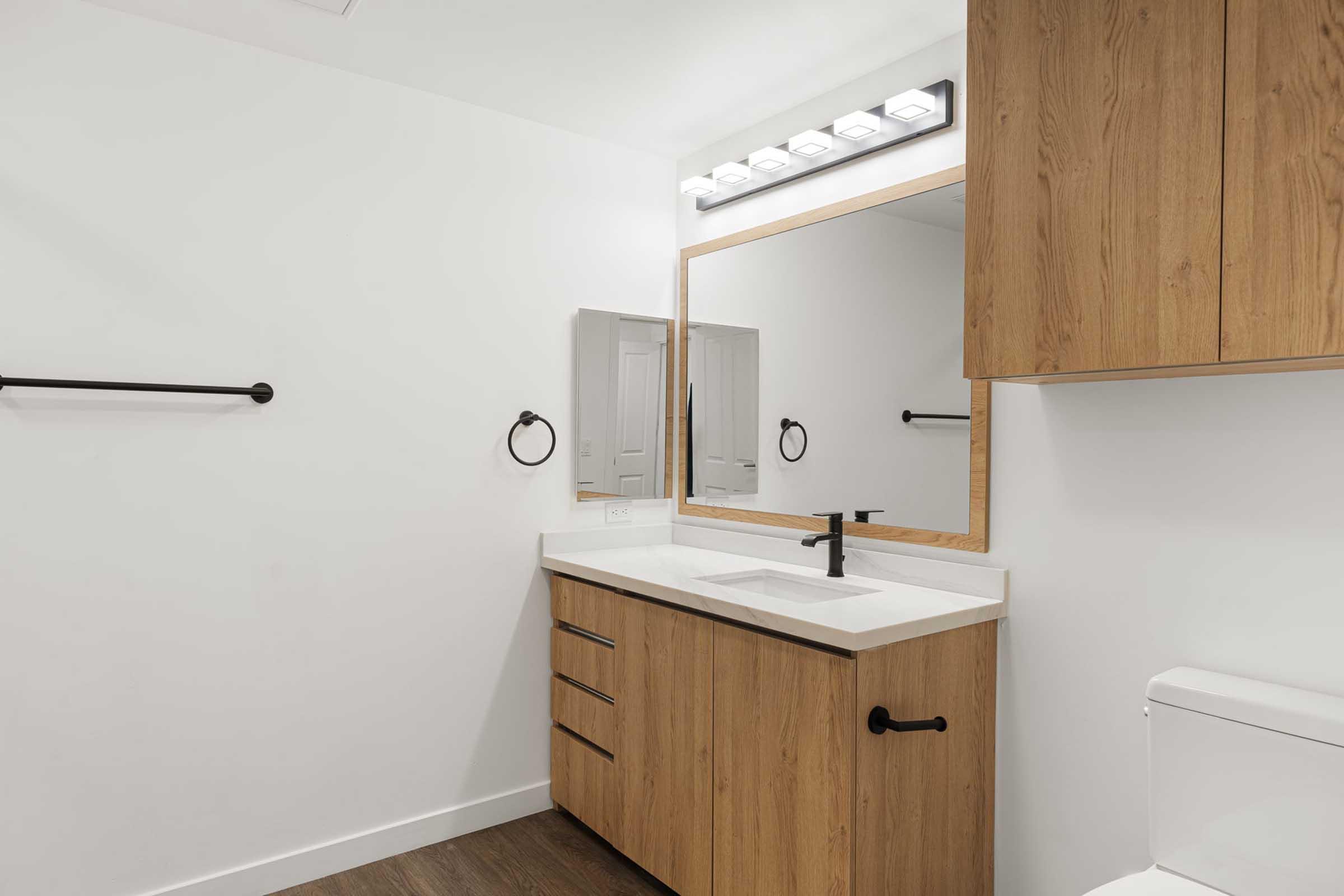 Modern bathroom featuring a wooden vanity with a white countertop and a sleek black faucet. A large mirror is above the sink, flanked by bright LED lights. The walls are painted white, and there is a towel bar and toilet in the space. The flooring is dark wood, providing a contemporary aesthetic.