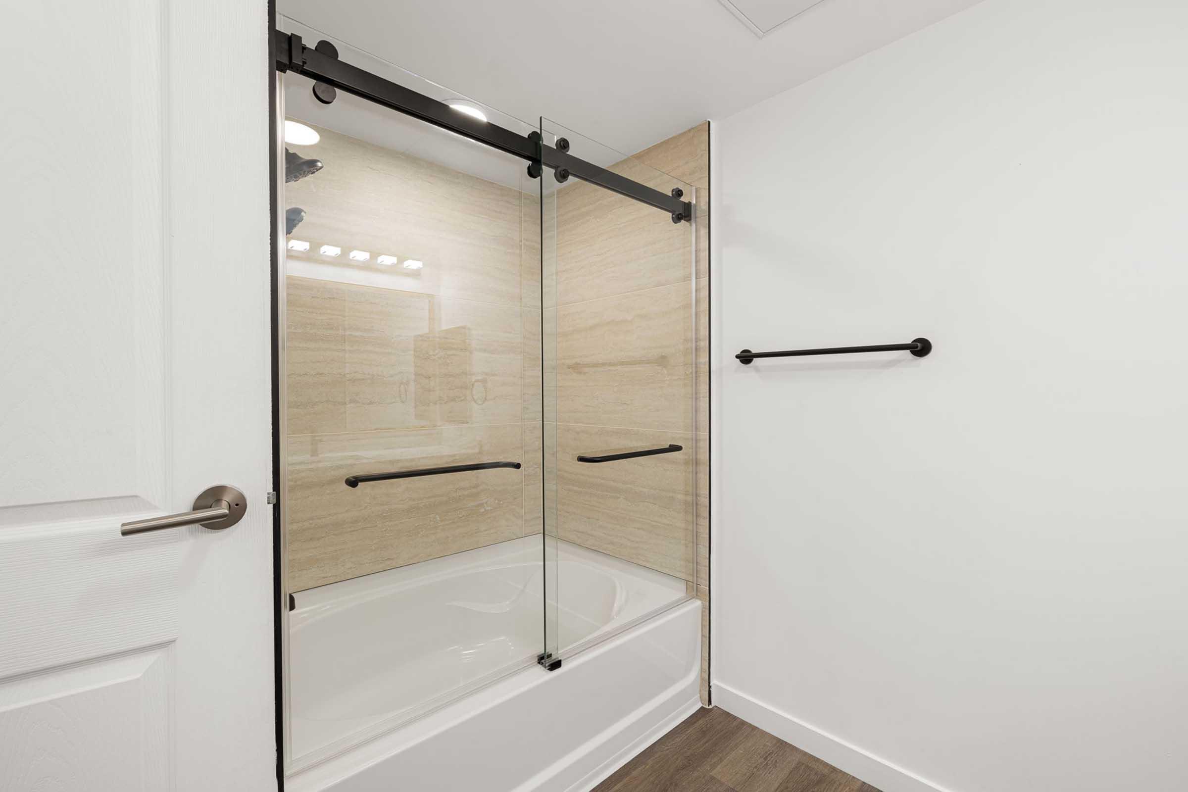 A modern bathroom featuring a glass shower enclosure with black sliding doors, a bathtub underneath, and beige tiled walls. There is a black towel bar mounted on the wall, and the space has a clean, minimalistic design with white walls and a wooden floor.