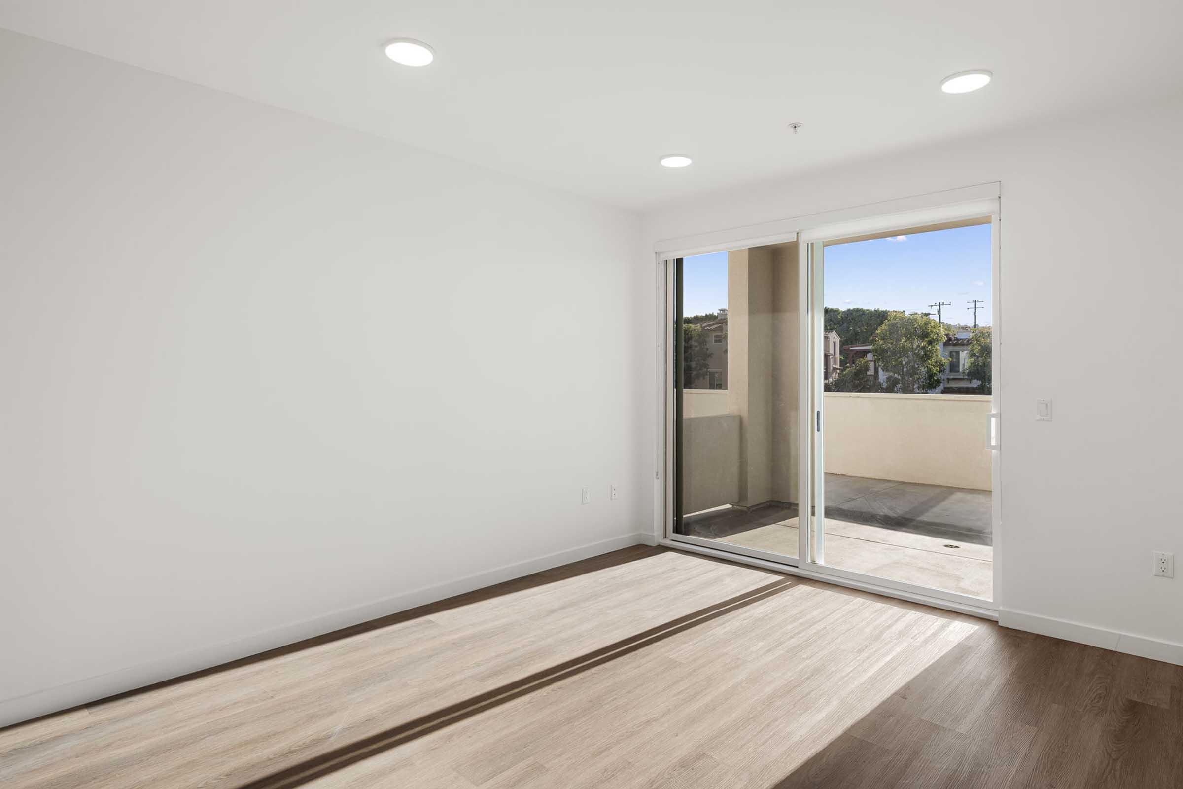Bright, empty room with large sliding glass door leading to a balcony. The walls are white, and there are recessed ceiling lights. Light-colored wooden flooring is visible, with sunlight streaming in from outside, hinting at a view of greenery beyond the balcony.