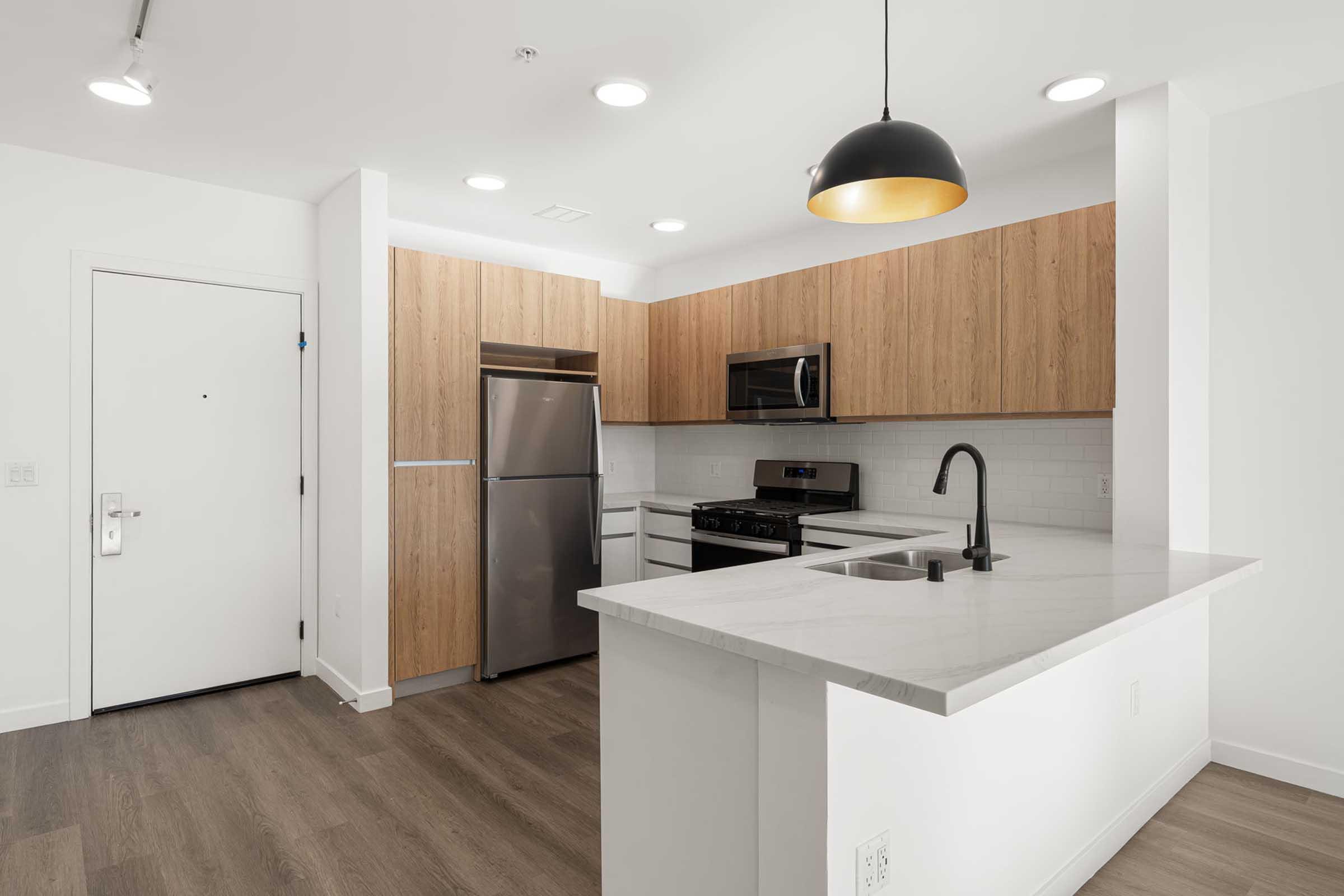Modern kitchen featuring a stainless steel refrigerator and microwave, a gas stove, and a sink with a sleek black faucet. The cabinetry is a light wood finish, complemented by a white countertop. Bright lighting illuminates the space, which has a white and wood color scheme, creating a clean and contemporary look.
