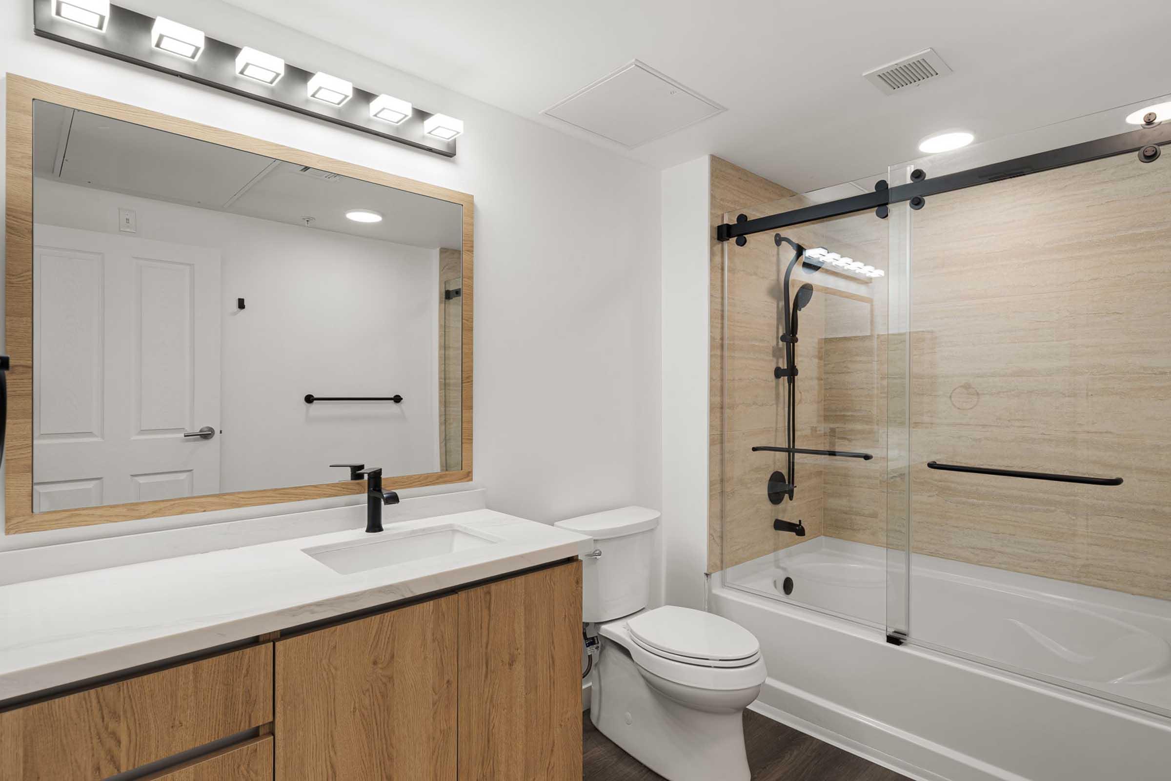 A modern bathroom featuring a large mirror above a white countertop with a sleek faucet. The space has wooden cabinetry, a glass shower enclosure, and a bathtub. Bright lighting highlights the neutral-colored walls and warm wood accents, creating a clean and inviting atmosphere.