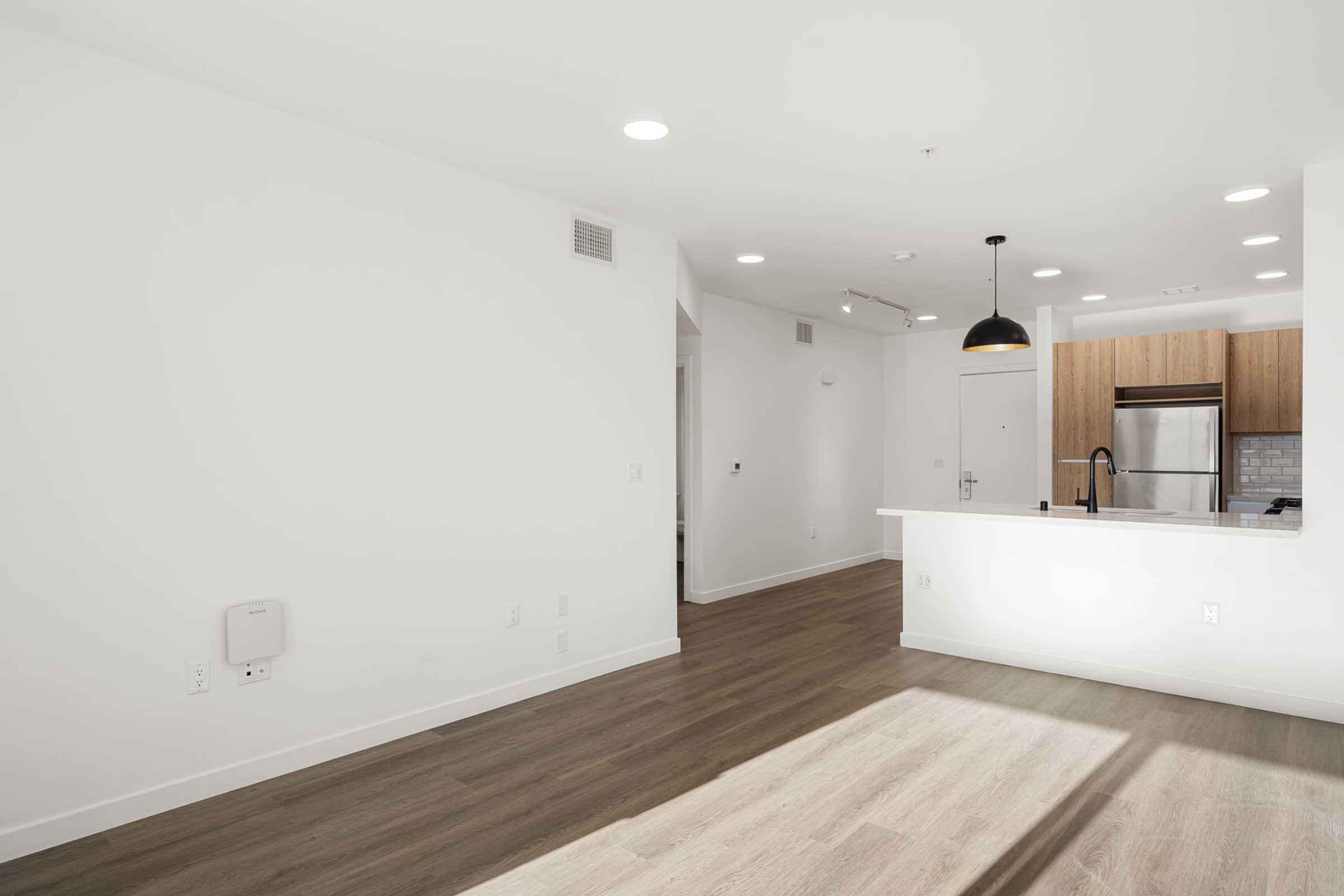 A modern, minimalistic interior of an apartment featuring a spacious living area with light wooden flooring, white walls, and a stylish kitchen with wooden cabinets and stainless steel appliances. Natural light streams in, creating a bright atmosphere.