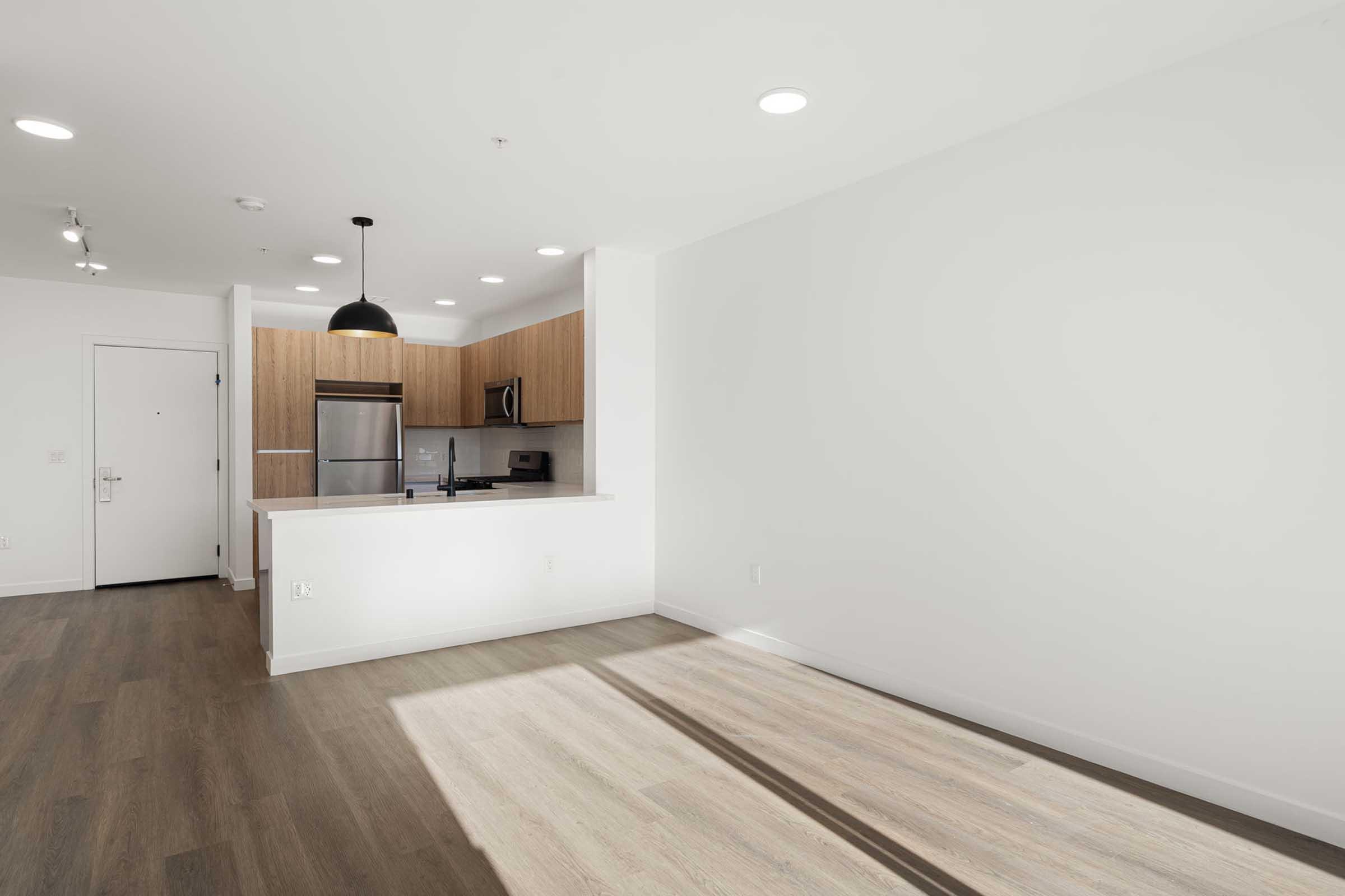 A modern, empty apartment interior featuring a light-filled space with wooden flooring. The kitchen is visible in the background, showcasing sleek cabinets, stainless steel appliances, and a black pendant light. The walls are painted white, contributing to a bright, airy atmosphere.