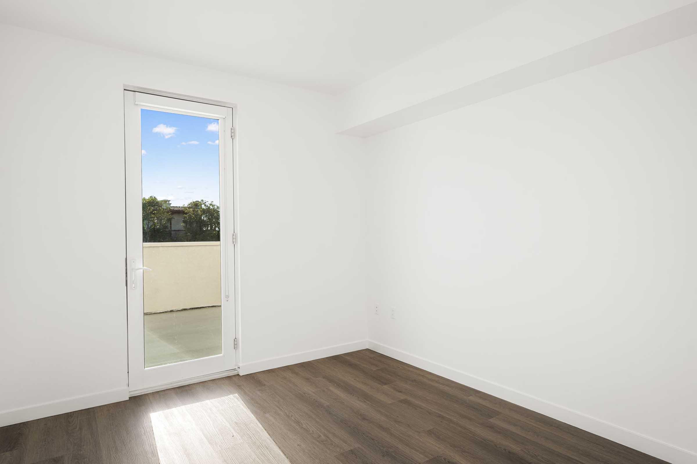 A bright, empty room with white walls and light hardwood flooring. There is a large window with a glass door leading to a balcony, allowing natural light to fill the space. The view outside includes a clear blue sky with some greenery visible. The room is devoid of furniture, offering a clean and minimalist appearance.