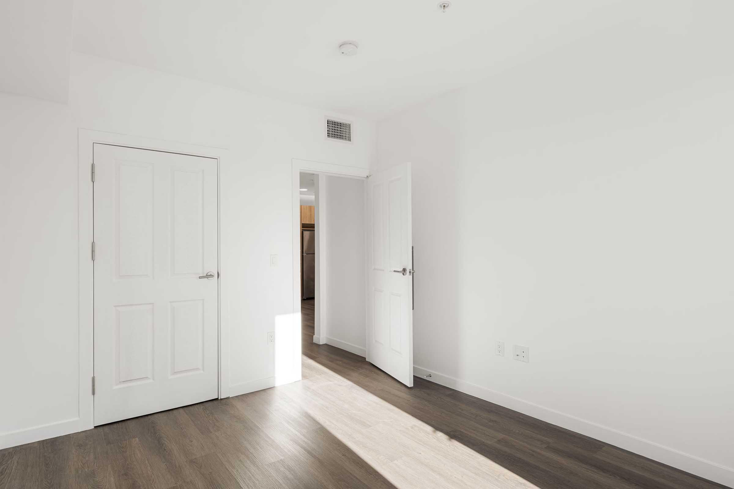 A bright, empty room with white walls and hardwood floors. There are two doors: one closed and one partially open, leading to another space. Natural light streams in, creating a clean and minimalist atmosphere.