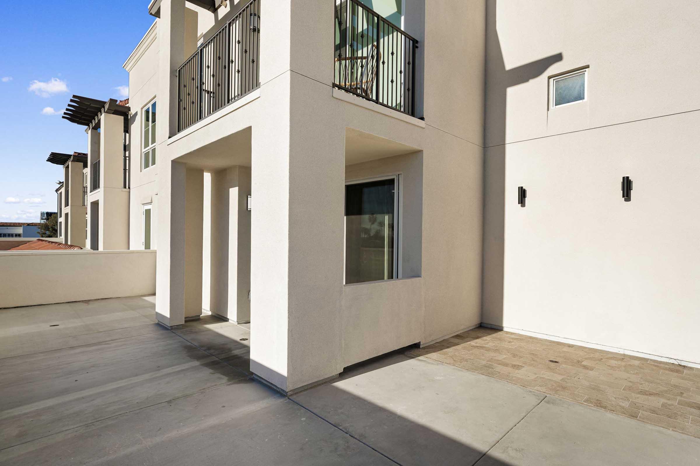 Exterior view of a modern building with a spacious balcony and concrete patio. The design features light-colored walls and black railings, under a clear blue sky. The area is well-lit with natural sunlight, showcasing the contemporary architectural style.