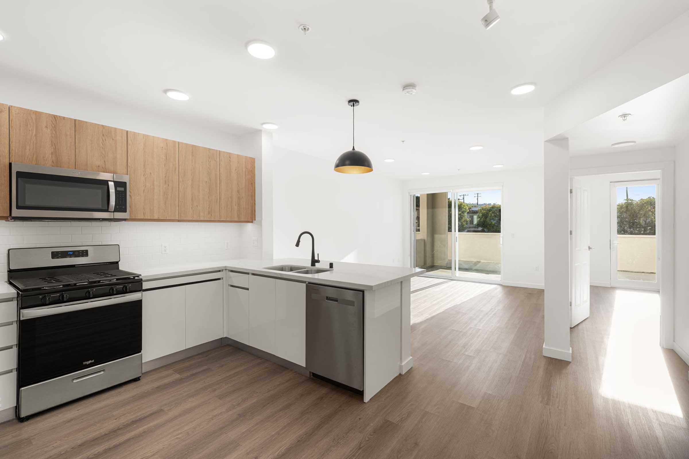 Modern kitchen featuring a combination of wood and stainless steel cabinetry, a gas stove, and a sleek sink. The layout includes an open space leading to a bright living area with sliding glass doors opening to a balcony. Light walls and hardwood flooring create a spacious atmosphere.