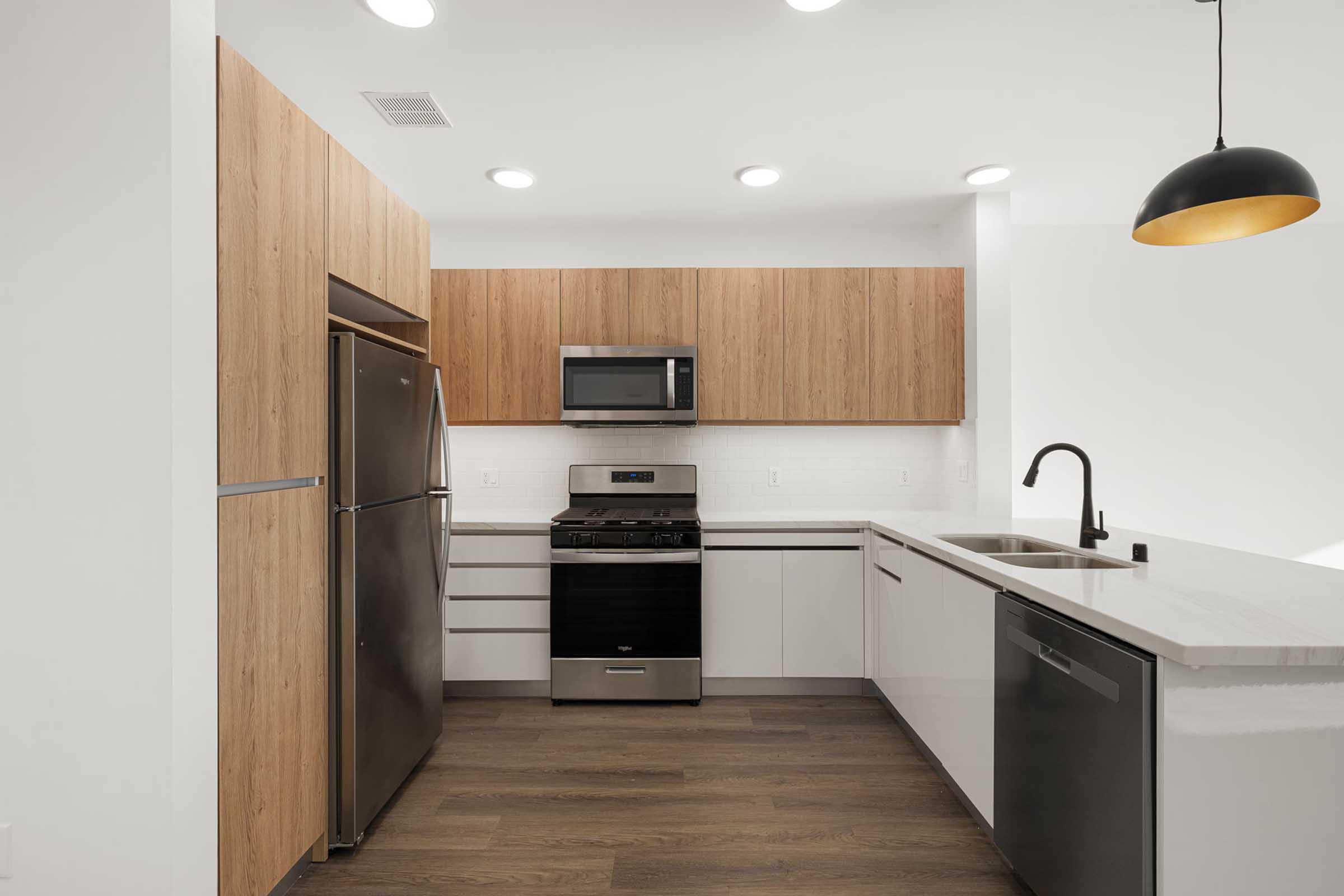 Modern kitchen featuring stainless steel appliances, including a fridge, microwave, and stove. The cabinetry is a mix of wood and white finishes, with a sleek sink and faucet. The space has warm wooden flooring and overhead lighting, creating a bright and inviting atmosphere.