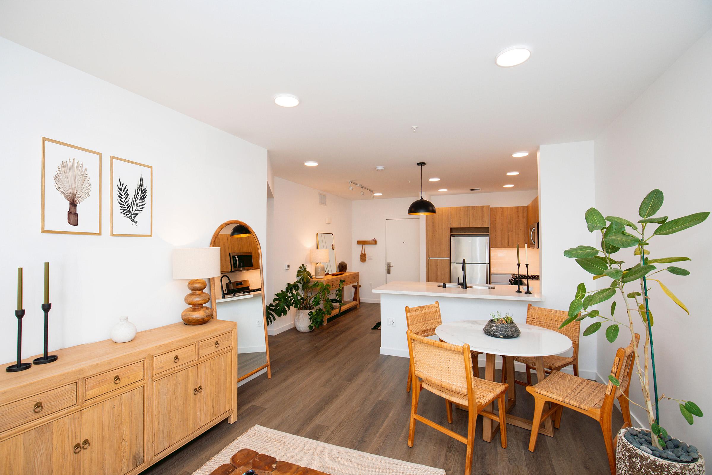 Modern living room and kitchen area featuring wooden furniture, light walls, and a round dining table with wicker chairs. Decorative plant in a pot, framed nature art on the wall, and stylish lighting create a cozy atmosphere. The space is well-lit with natural light and contemporary decor.