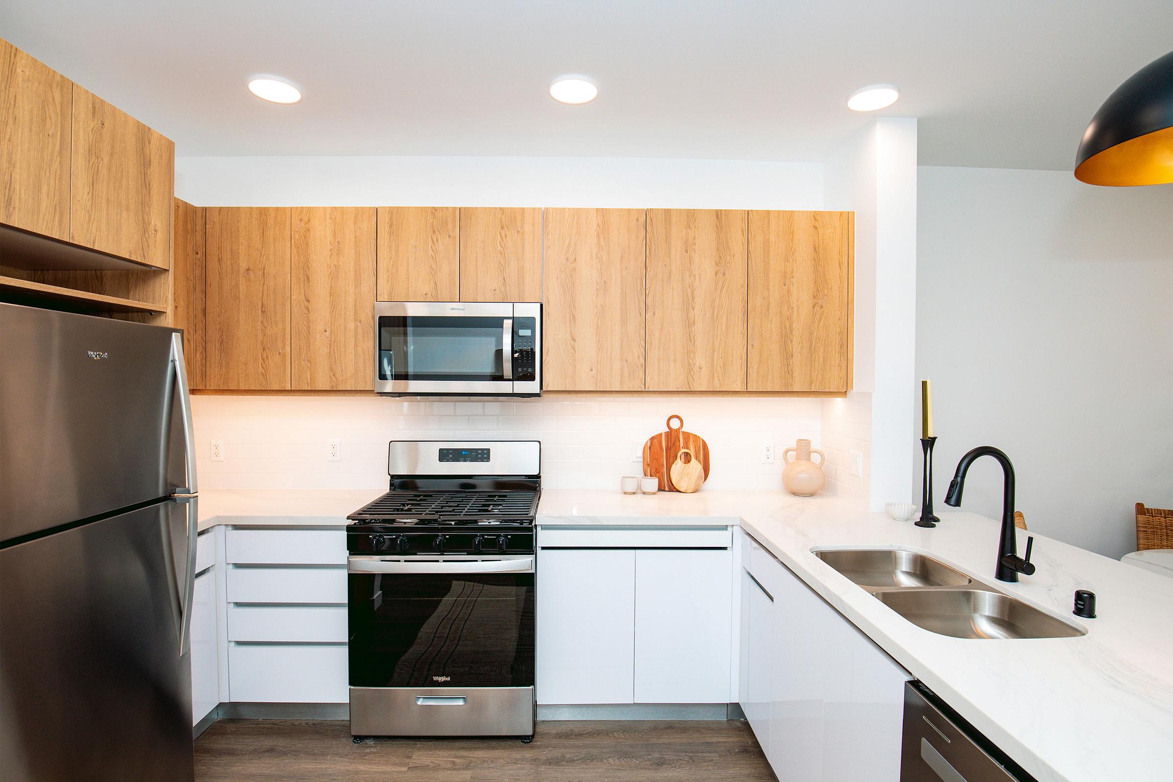 Modern kitchen featuring a stainless steel refrigerator and an oven with a gas range. The cabinetry has a mix of wood and white finishes. A sink with a black faucet is positioned near a stylish cutting board and decorative items on the counter. Bright lighting and a minimalist design create a clean and inviting space.