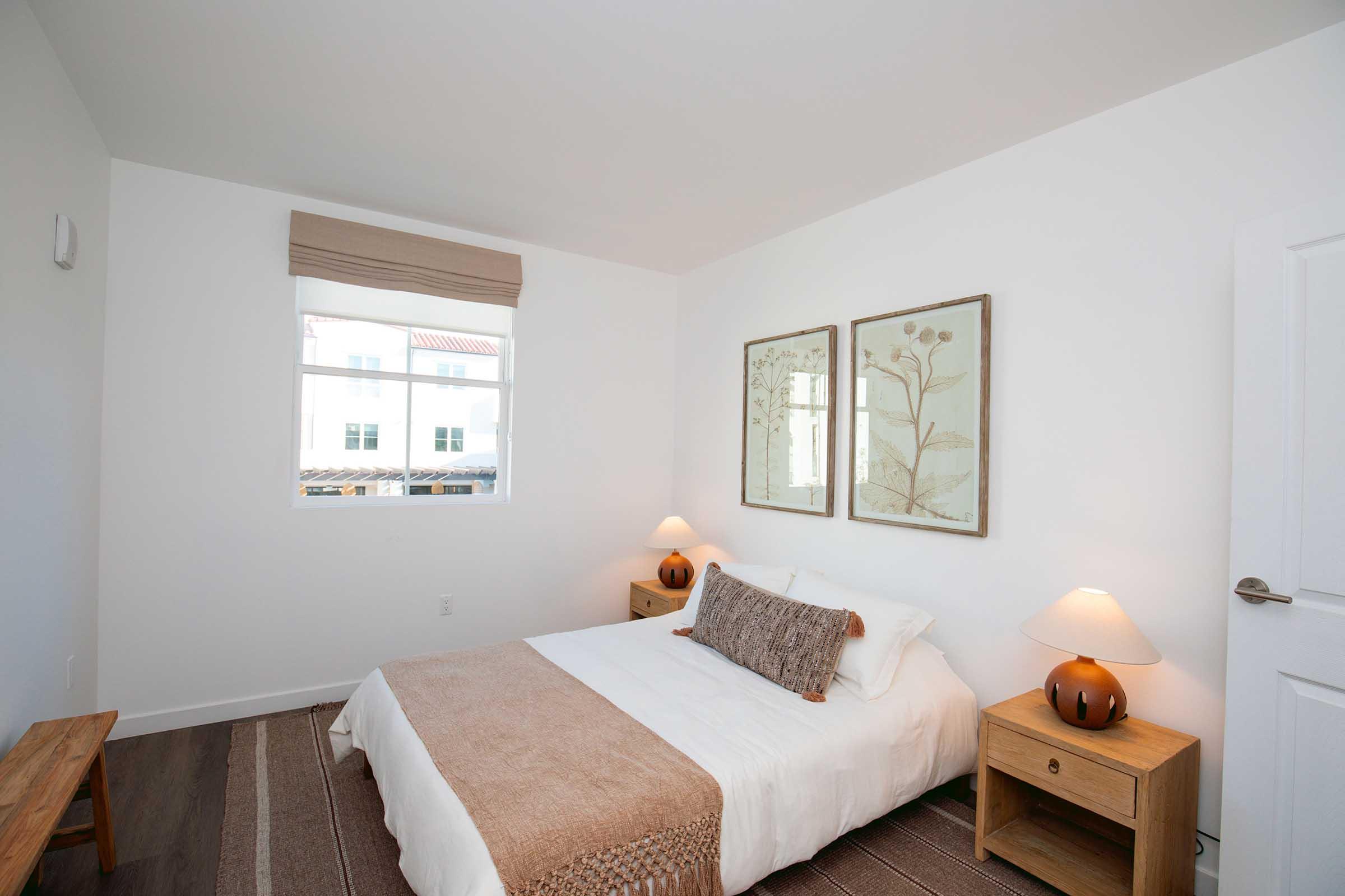 Cozy bedroom featuring a neutral color palette. A double bed with a light bedding set and a decorative throw sits between two wooden nightstands with lamps. Natural light filters through a window with a simple shade. Two framed botanical prints adorn the wall, adding a touch of nature to the serene space.