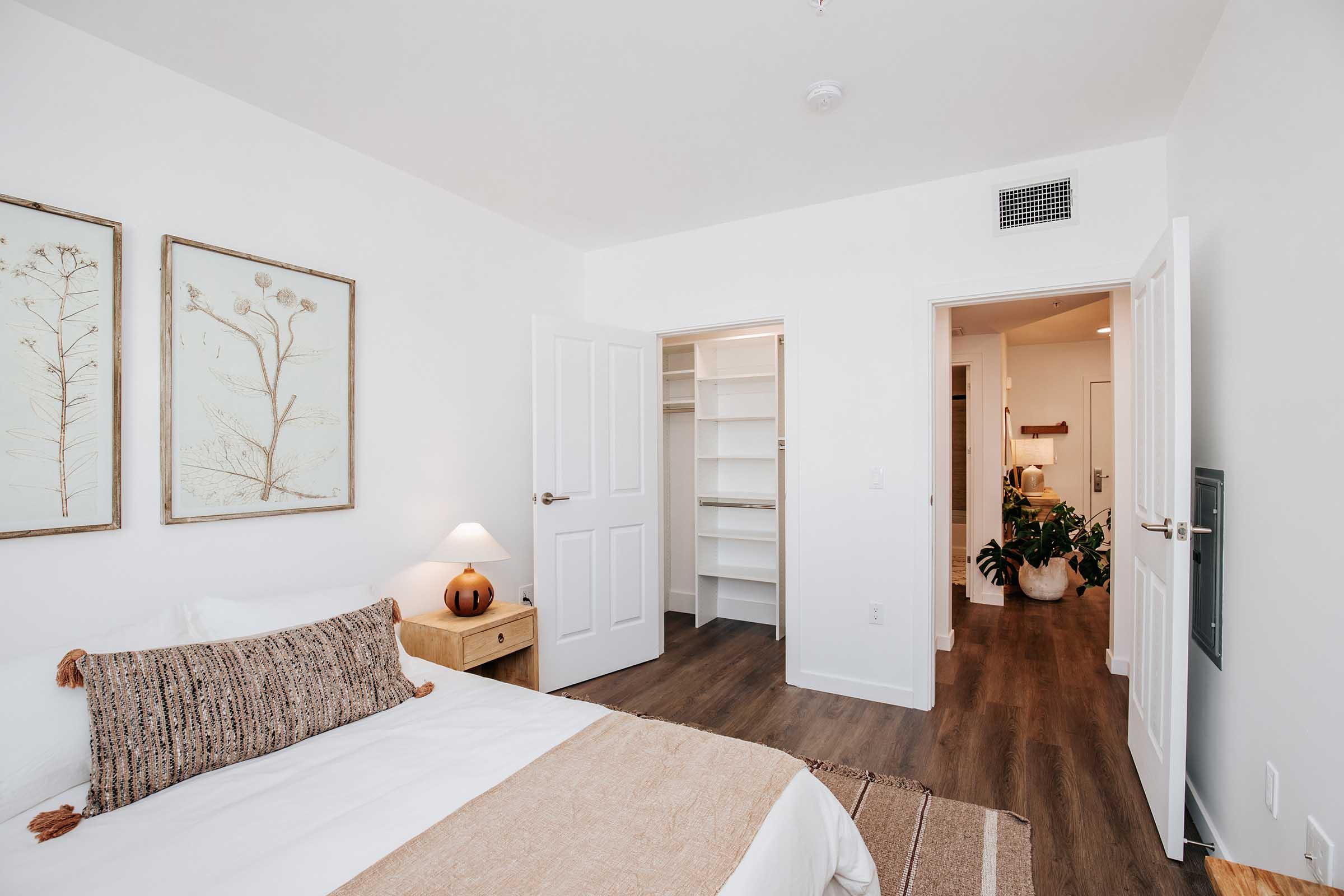 A cozy, modern bedroom featuring a large bed with decorative pillows and a throw. Two framed botanical prints adorn the wall above the bed. A nightstand with a lamp is beside the bed. In the background, a doorway leads to a closet with open shelving. A plant is visible in a pot near the closet entrance.