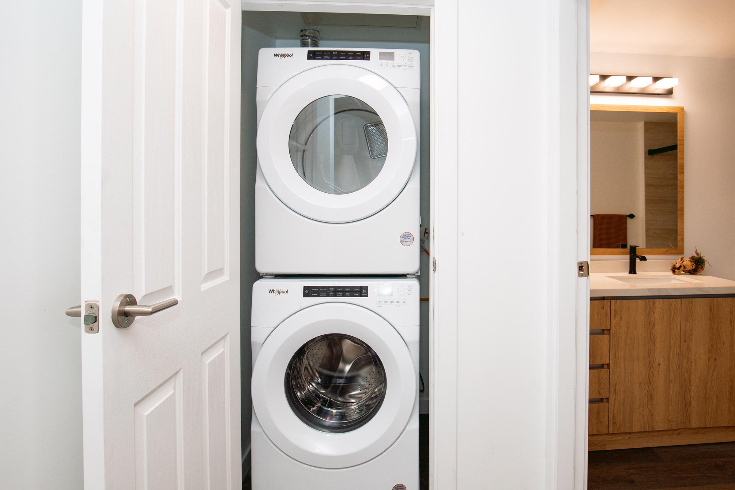 A compact laundry area featuring a stacked washer and dryer in white next to a bathroom. The doors are open, revealing the appliances. In the background, there is a bathroom vanity with a mirror and light fixtures, along with wooden cabinetry.
