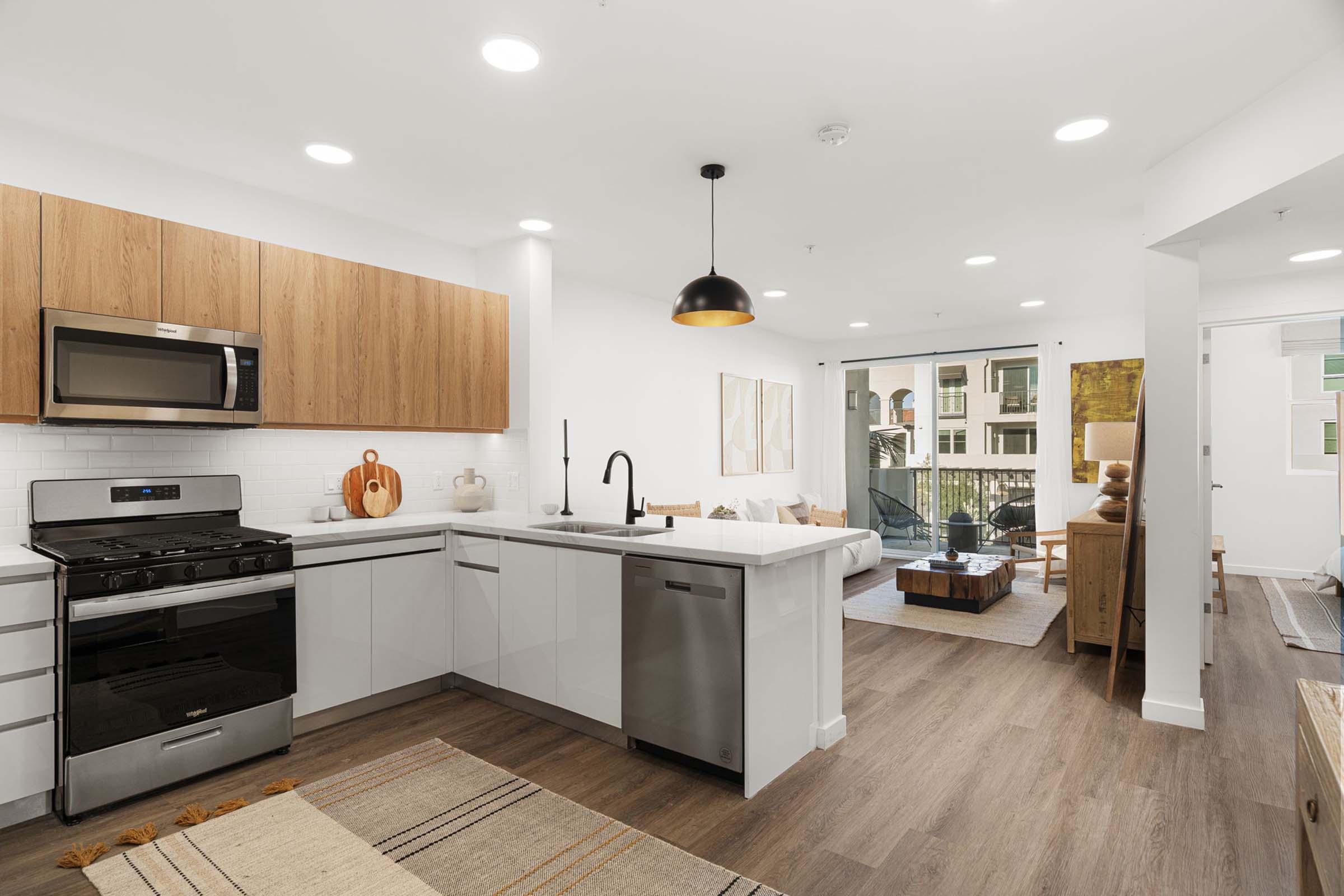 Modern kitchen and living area featuring stainless steel appliances, wooden cabinetry, and a bright, open layout. A dining table is visible along with comfortable seating, decorative elements, and natural light streaming through large windows. The space has a contemporary aesthetic with neutral tones.