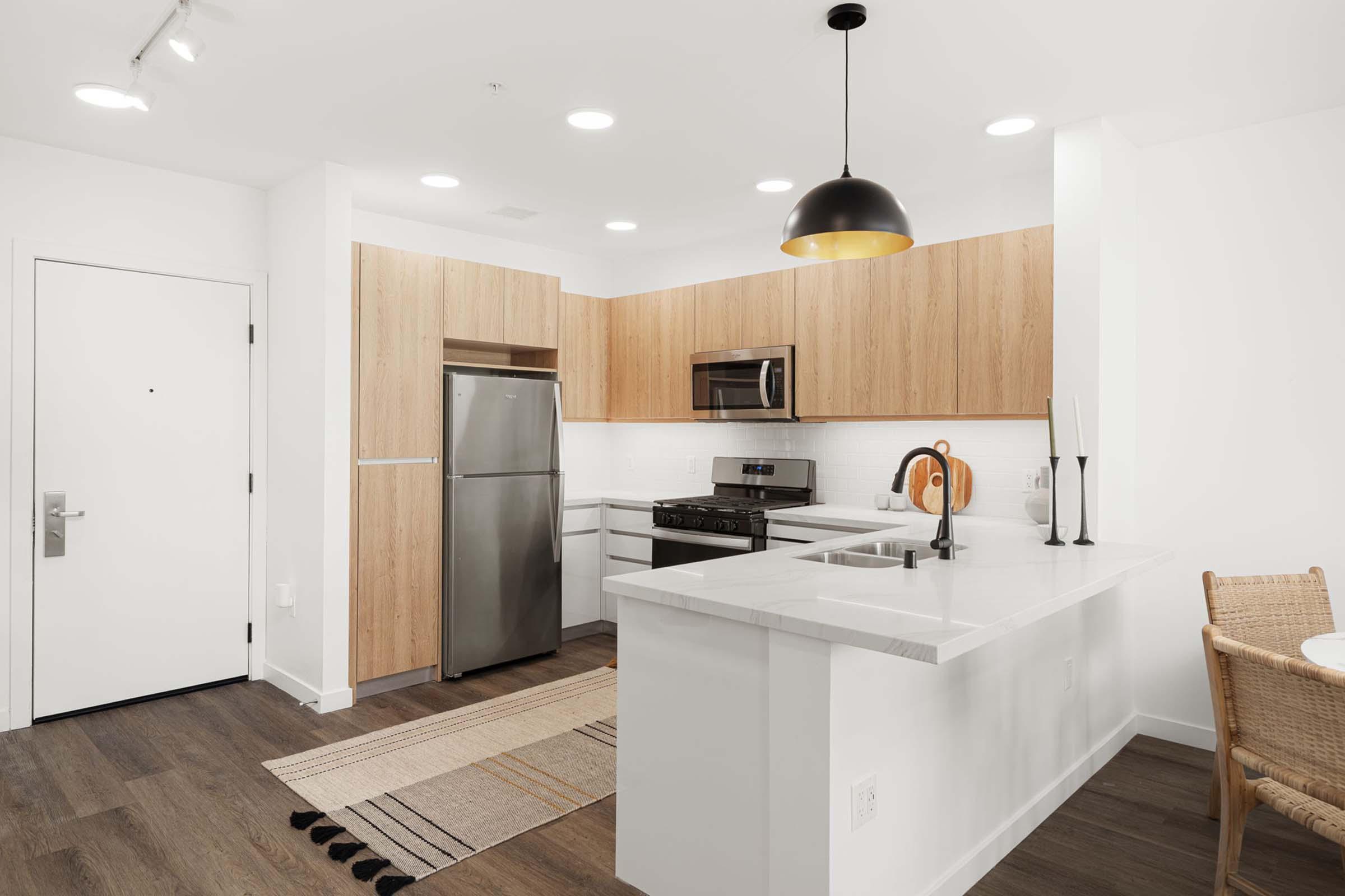 Modern kitchen featuring stainless steel appliances, including a refrigerator, stove, and microwave. The cabinetry is light wood, and the countertops are white. There's a small dining area with a wicker chair and a decorative rug. Bright lighting enhances the open, inviting atmosphere.