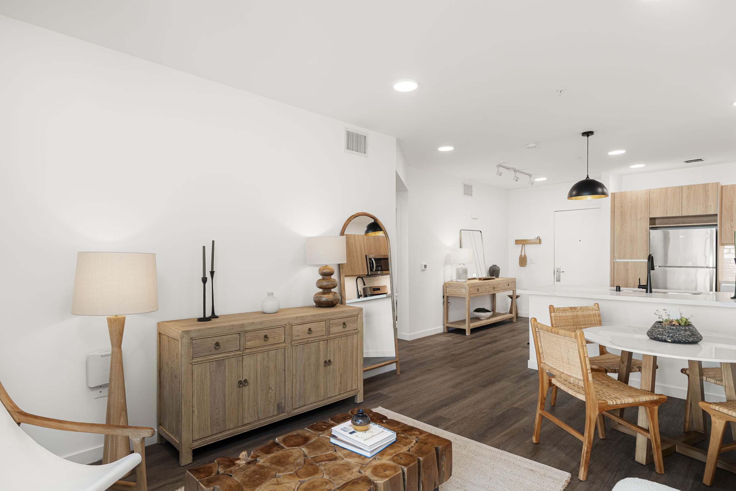 Modern living room and dining area featuring a light color palette. A wooden sideboard and round dining table with woven chairs are visible, along with a decorative lamp and a stylish light fixture. The space has an open layout with simple decor, giving a clean and inviting feel.