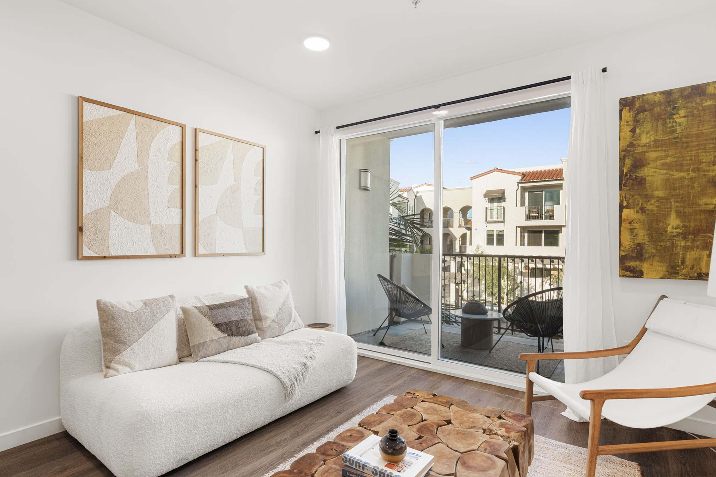 A modern living room featuring a cozy white sofa with decorative pillows, two abstract framed artworks on the wall, and a large sliding glass door leading to a balcony. There are two outdoor chairs on the balcony and a stylish wooden coffee table in front of the sofa, creating a welcoming atmosphere.