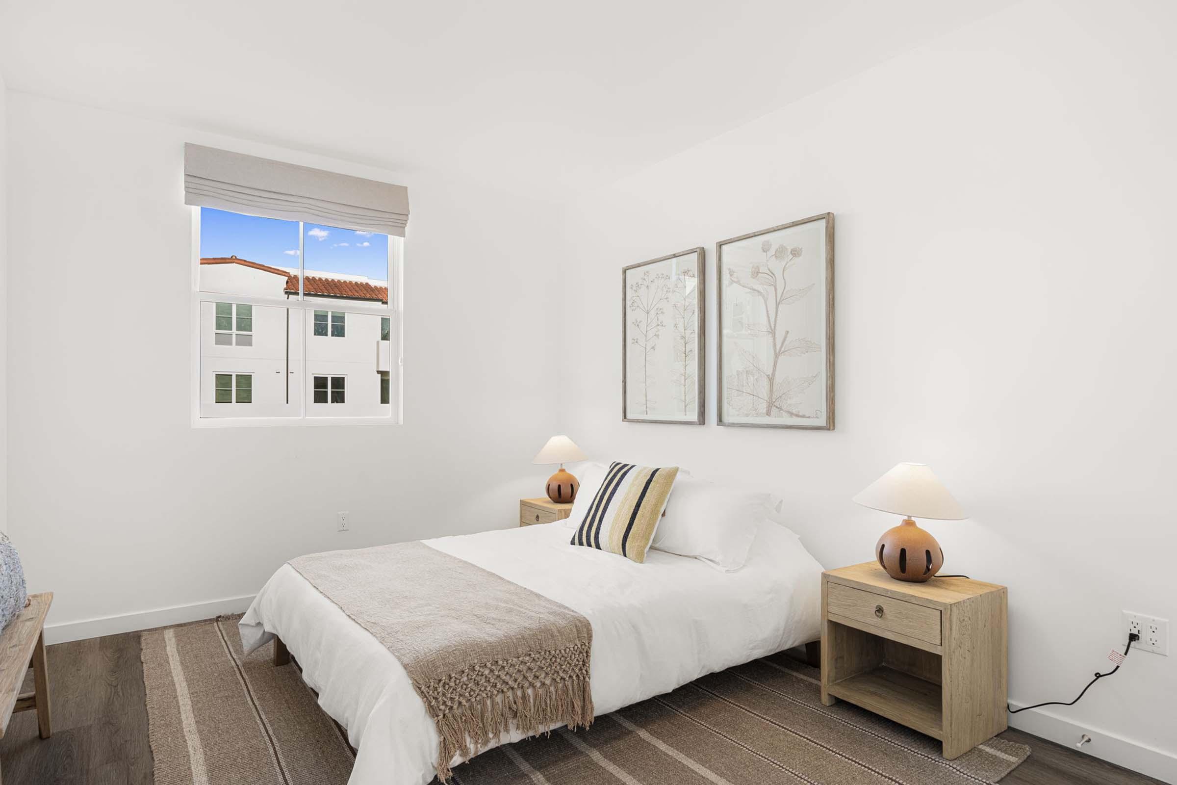 Bright and airy bedroom featuring a white bedspread, two bedside tables with lamps, and framed botanical art on the wall. A window reveals a blue sky and buildings outside. The floor is adorned with a striped area rug, creating a cozy atmosphere.