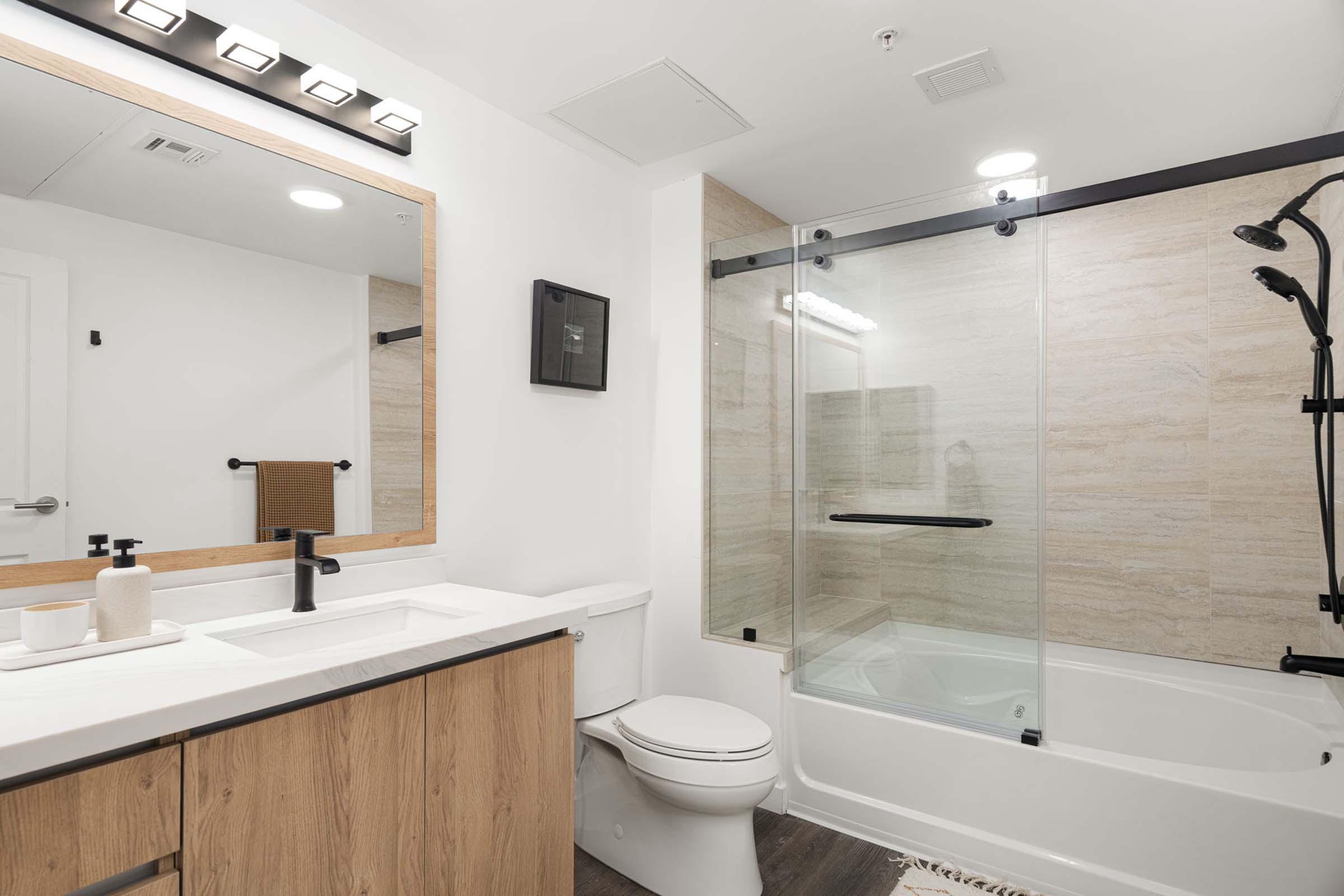 A modern bathroom featuring a glass shower enclosure, a white bathtub, and a sleek vanity with a large mirror. The cabinetry is light wood, and there are minimalist decor elements, including towel racks and toiletries. A bright light fixture is mounted above the mirror, enhancing the clean, contemporary aesthetic.