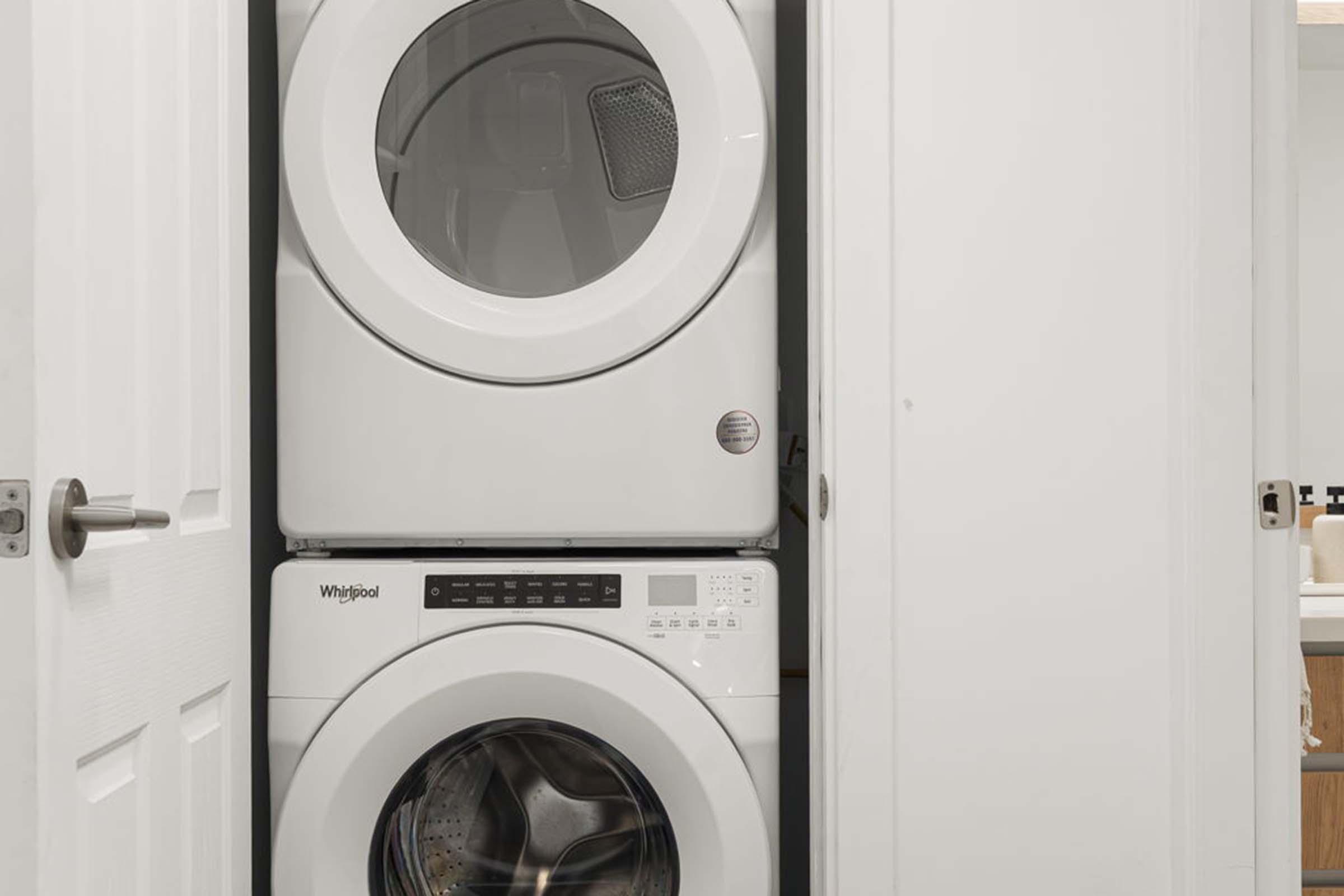 A compact laundry area featuring a stacked washer and dryer. Both appliances are white with a modern design. The washer is located on the bottom, while the dryer sits above it. The space is enclosed by white cabinetry and a door is visible, indicating a tidy and efficient use of space.