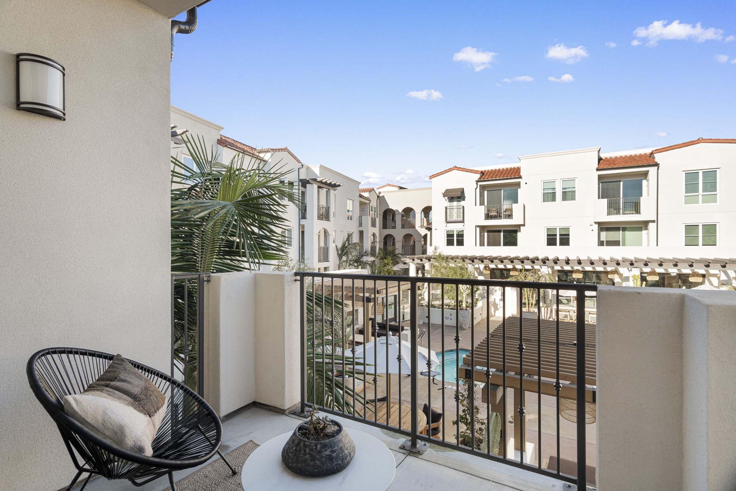 A view from a balcony overlooking a pool area of an apartment complex. The balcony features a round chair, a small table with a decorative item, and potted plants. Nearby buildings have a modern design with white walls and red-tiled roofs against a clear blue sky with a few clouds.