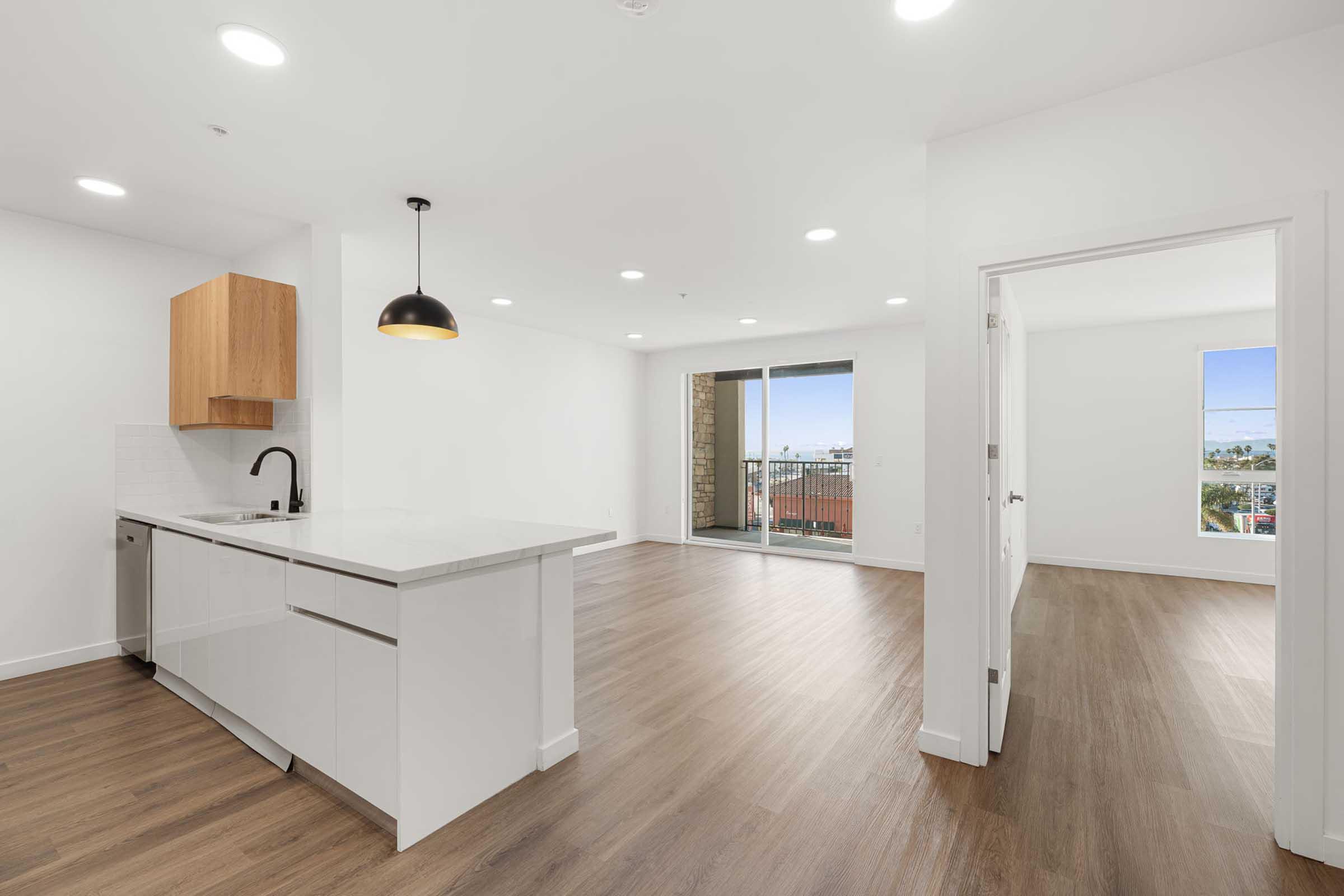Bright and modern open-concept living space featuring a sleek kitchen with a white countertop and wooden cabinets, hardwood floors, recessed lighting, and a large window with a view. The area connects to a balcony, providing natural light and a spacious atmosphere.